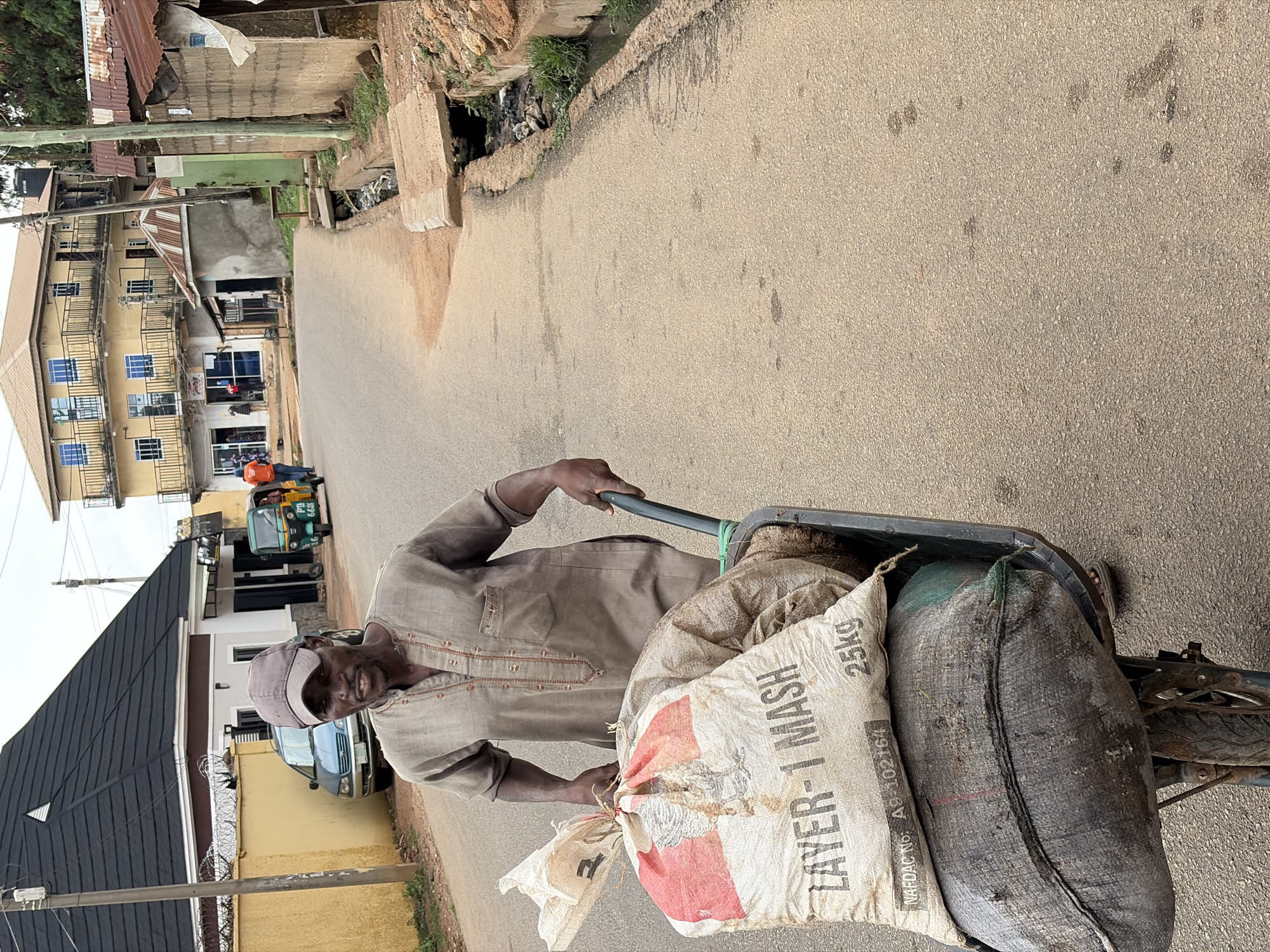 The image shows a man pushing a loaded cart down a street in Jos, Nigeria, during daytime. The man, with dark skin, a short beard, and a light-colored cap, wears a light brown, long-sleeved shirt with embroidery. He smiles at the viewer while pushing a single-wheeled cart, which appears to be a wheelbarrow, filled with sacks.
The most prominent sack in the cart is white with red and black lettering, clearly displaying "LAYER-1 MASH", "25kg", and "NAFDAC No: A8-102164". A second, dark grey/black sack is also visible in the cart.
The street surface is an asphalt or gravel road, showing signs of wear and dirt. On the left side of the street, there is a multi-story yellow building with dark metal balconies and numerous windows, along with other residential or commercial structures, some featuring corrugated metal roofs. A three-wheeled vehicle (tricycle/keke Napep) and other people are visible further down the street. A blue car is parked near a building with a dark roof and a fence topped with barbed wire. Utility poles stand alongside the road. The scene depicts a lively, everyday urban environment.