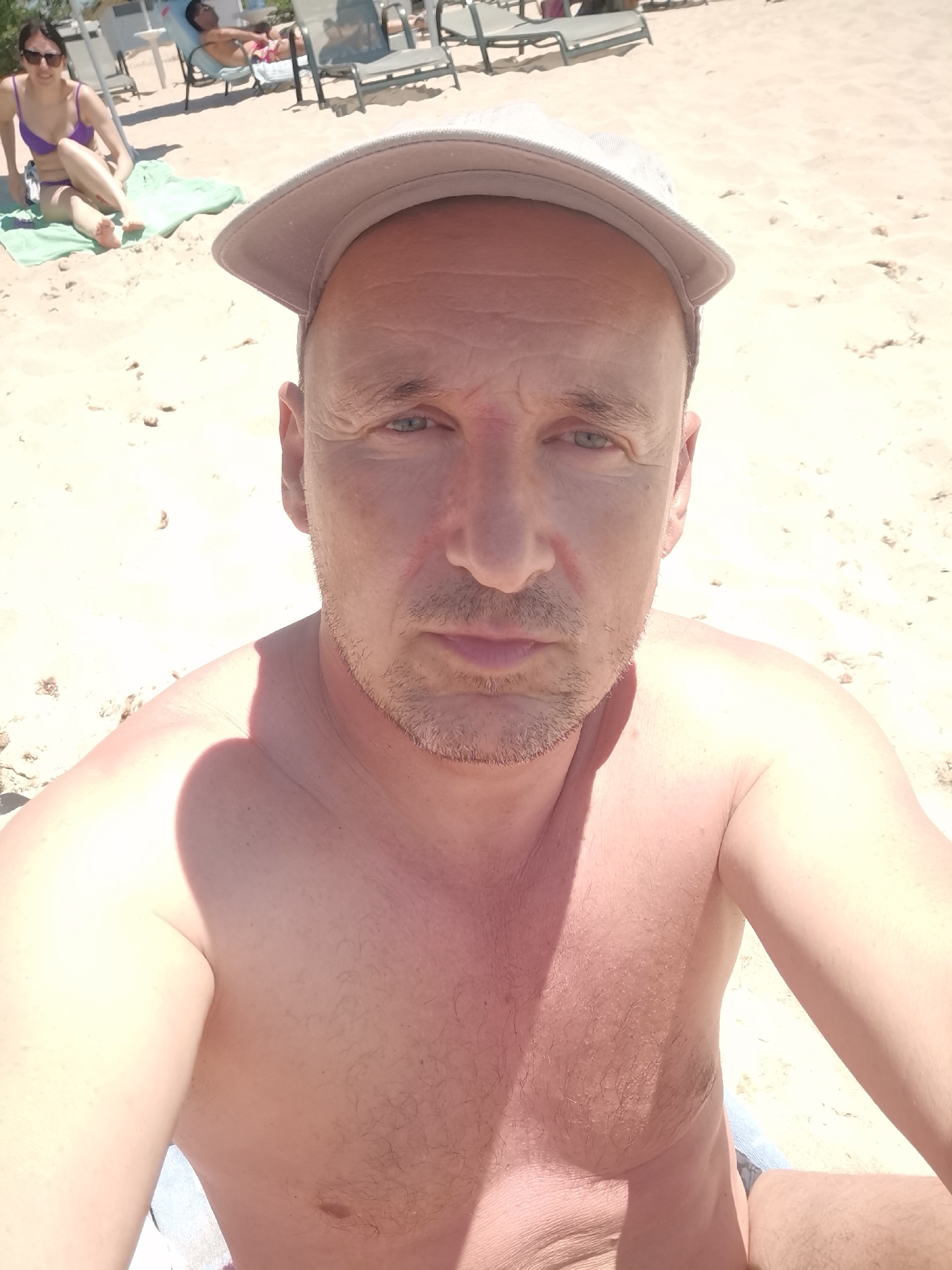 A man is taking a selfie on a sunny beach in Kranevo, Bulgaria. He is wearing a light-colored baseball cap and appears to be topless. The background shows a sandy beach with a few lounge chairs and two women in the distance. One woman, wearing a purple bikini, is sitting on a towel, and another person is lying on a lounge chair. The man's face is partially illuminated by sunlight, casting a shadow on his left side. The overall scene depicts a casual day at the beach.