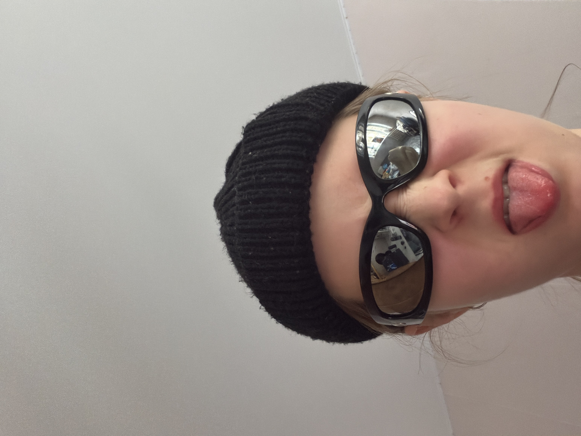A close-up, slightly rotated portrait captures a person with a playful expression, sticking their tongue out. The subject's head is tilted to the right, and they are wearing a dark, possibly black, knitted beanie and prominent black sunglasses with highly reflective, mirrored lenses. The sunglasses obscure the eyes entirely. The reflections in the lenses show an indoor environment, with hints of light-colored walls or ceilings and some indistinct indoor structures or furniture, suggesting a room or vehicle interior. The person's skin tone is fair, and a hint of a smile is visible around the mouth, contributing to the lighthearted mood. The background is a plain, undifferentiated light gray surface, providing no specific location cues. There is no visible text in the image. The scene is located in Perm, Russia.