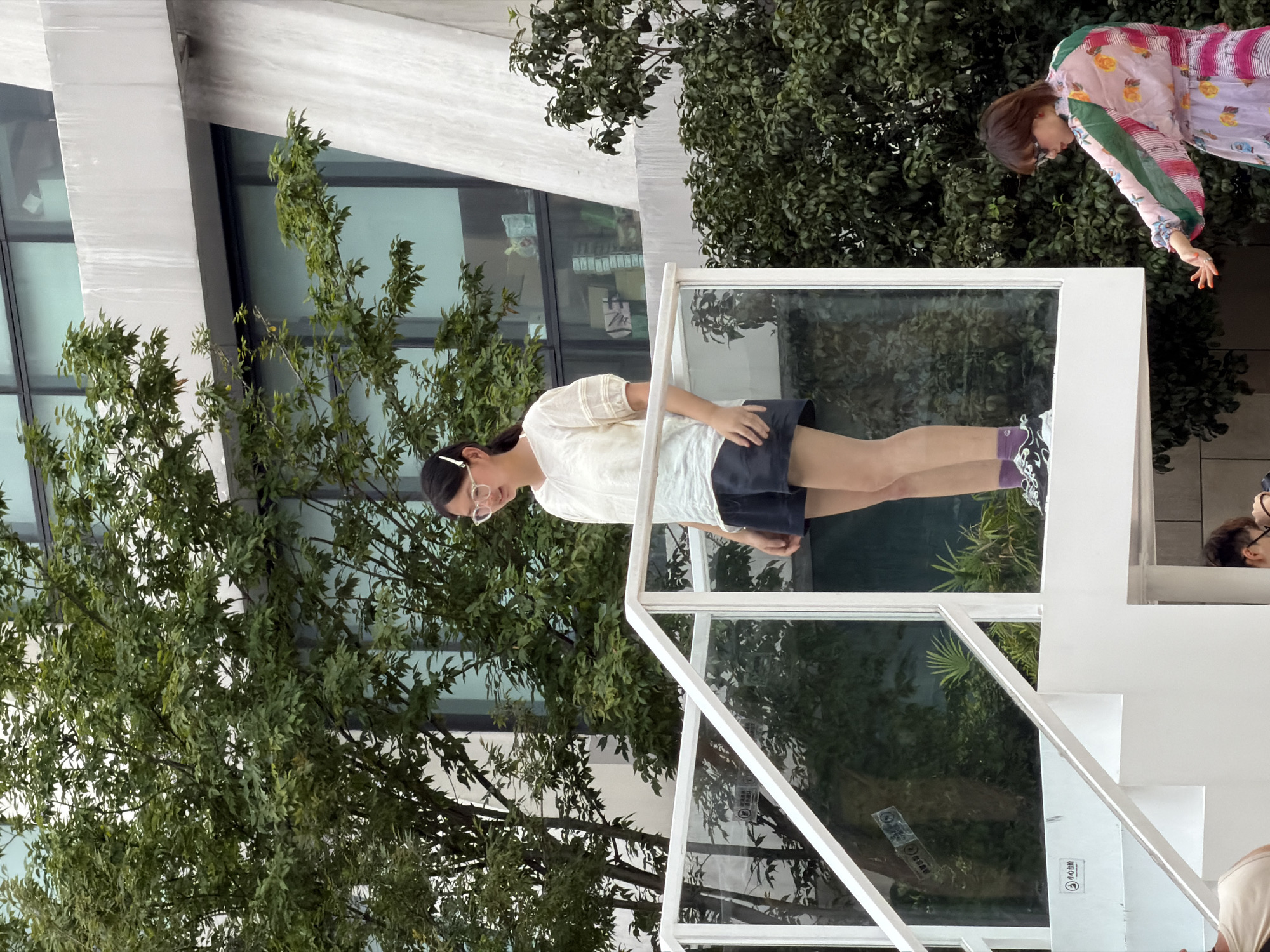 In Yuzhong District, China, a young woman wearing glasses, a white short-sleeved blouse, and dark shorts stands on a white elevated platform with a glass railing. She is looking downwards. To her right, another woman in a colorful floral dress is reaching out her left hand, her right hand also visible. The background features lush green trees and a modern building with large windows.