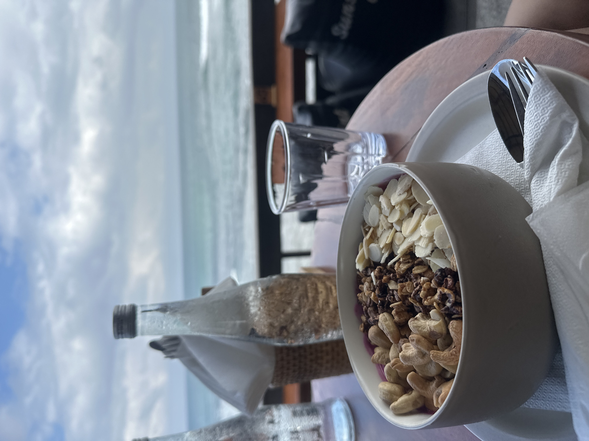 This media file captures a close-up of a breakfast bowl placed on a wooden table, with a serene ocean backdrop. The bowl is filled with granola, slivered almonds, and cashews, suggesting a healthy and hearty meal. To the right of the bowl, a white napkin is neatly folded, holding a spoon and fork, indicating that the meal is ready to be eaten.
In the background, a glass of water sits on the table, and a glass bottle, possibly containing water or a beverage, is also visible. The ocean stretches out beyond, with a slightly overcast sky suggesting it might be morning or late afternoon. The gentle waves and the vast expanse of water create a tranquil and relaxing atmosphere.
The setting appears to be an outdoor dining area, perhaps a cafe or restaurant, overlooking the beach. The warm tones of the wooden table contrast with the cool blues of the ocean and sky. There is no discernible human presence in the frame, emphasizing the peacefulness of the scene. The overall impression is one of a calm and enjoyable start to the day, or a relaxing moment by the sea.
The phrase "Sun S..." is partially visible on a black object in the background, but it's too obscured to read fully. The location context of North Kuta, Indonesia, further enhances the image's tropical and coastal ambiance.