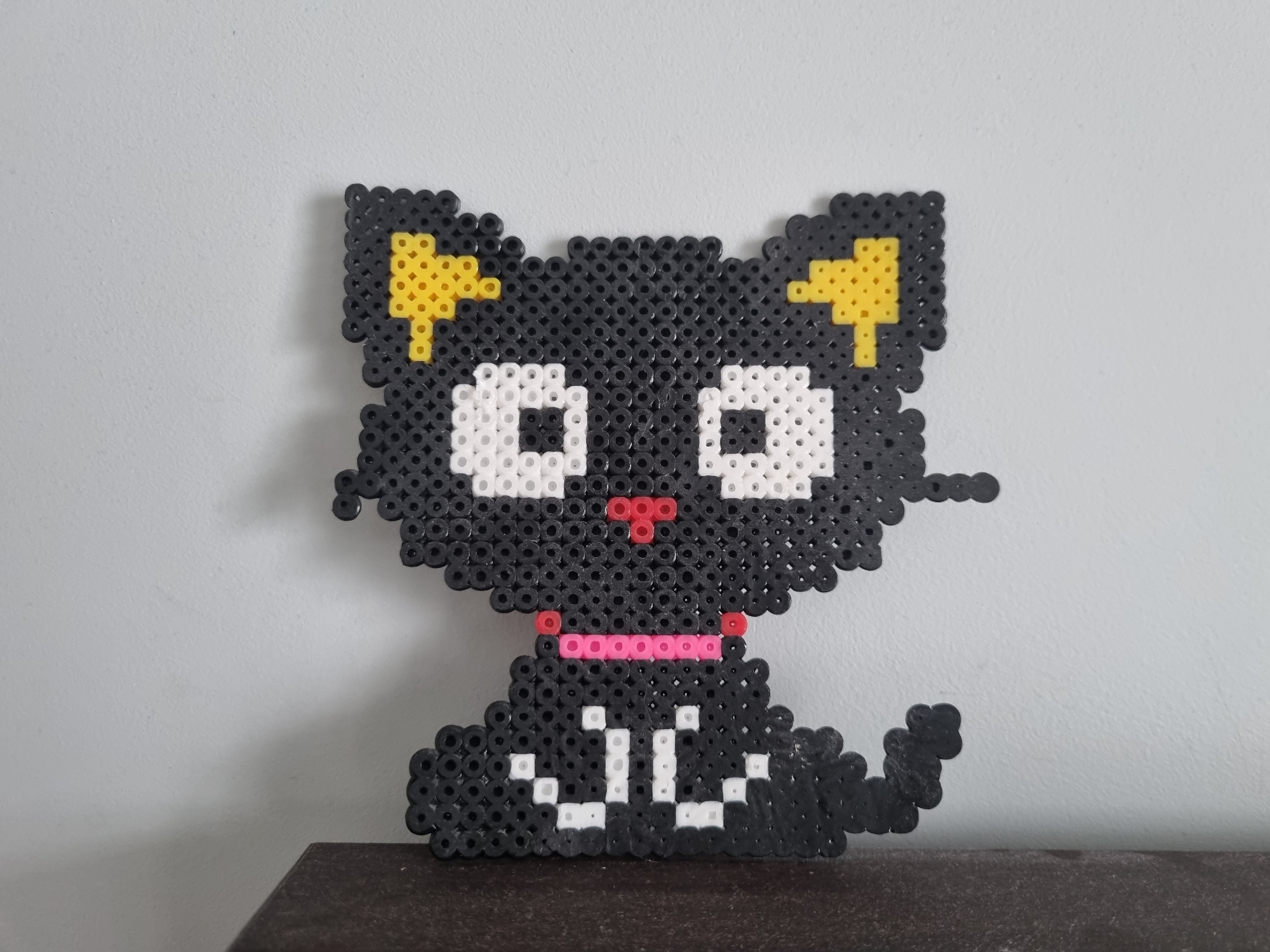 A black cat figurine made of fuse beads rests on a dark brown surface, possibly a table or shelf, in front of a light gray wall. The cat features white eyes, a pink nose and collar, yellow ear markings, and white markings on its chest. The background is a plain, light gray wall with no other visible details. The composition is a close-up shot, focusing primarily on the cat figurine. The city of Khimki, Russia is not discernible from this image.
