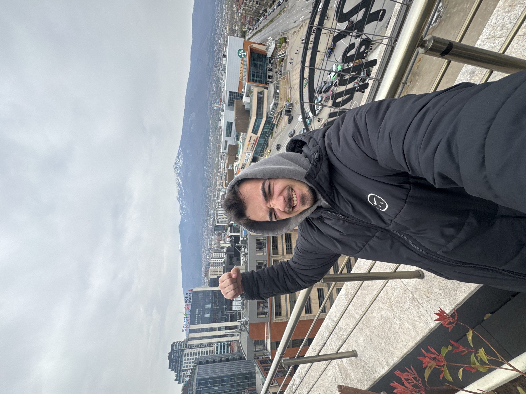 A medium shot shows a man smiling with a raised fist, leaning against a metal railing, with an overview of Sofia, Bulgaria, in the background. The man wears a gray hooded sweatshirt under a black puffer jacket with a white logo. The cityscape in the background includes tall buildings, a large shopping mall with a prominent "Pepsi" logo, and mountains in the distance. The sky is overcast, and the overall setting appears to be an urban environment on a cloudy day. Red flowers are visible in the lower right corner. The photo appears to be taken from an elevated position, such as a balcony or rooftop.