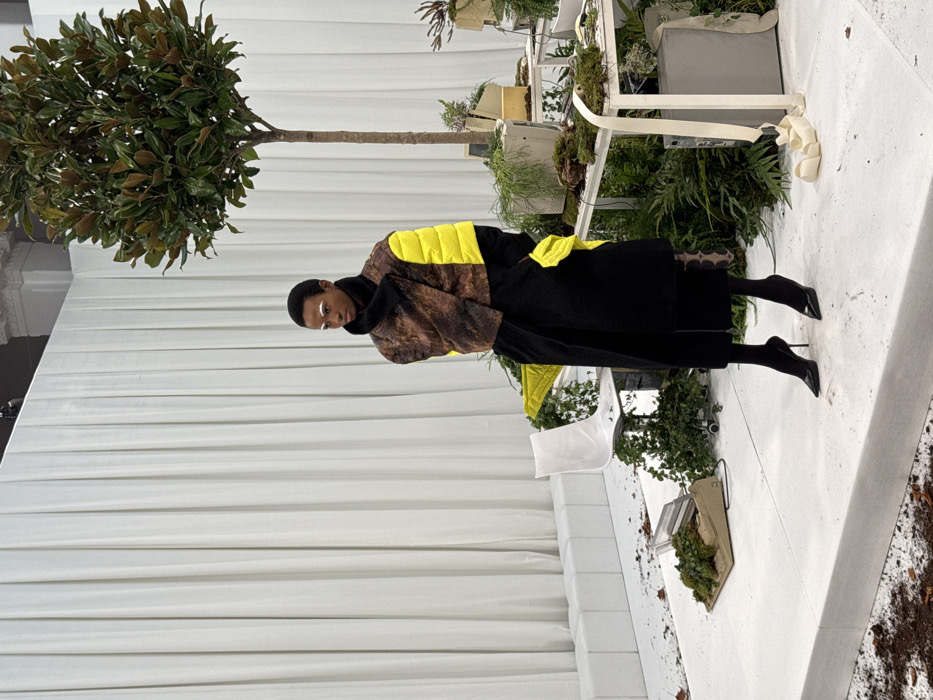 An interior scene, likely a fashion presentation, takes place within a building featuring classical architectural elements (fluted pilaster, cornice) in Milan, Italy. A single Black individual, presenting as a model, stands centrally on a raised white platform, oriented slightly left. The individual wears a high-collared black underlayer, a cropped, sleeveless outer jacket with distressed brown and bright yellow padded sections, a long black overcoat, black trousers, and black high-heeled pumps. Their short dark hair is visible, and their eyebrows and upper eyelids are marked with distinct white makeup.
The background consists of a tall, pleated white curtain extending across the width of the frame, with architectural details visible above it. The white floor platforms are sparsely littered with dark organic material, resembling soil or dead leaves. The set includes a small tree with dark green foliage, centered on a white platform to the right of the model. Multiple white tables or display stands are arranged at varying heights, primarily to the right. These tables display vintage technology, such as CRT computer monitors and keyboards, extensively integrated with green moss, ferns, and vines. A white office chair with a metallic base is positioned on a lower platform to the left, also partially covered by green foliage. Further left, an obsolete tan-colored office machine, possibly a typewriter or cash register, rests on a low platform, partially obscured by moss. A long, draped white fabric ribbon extends from one of the tables to the floor on the right side of the frame.
