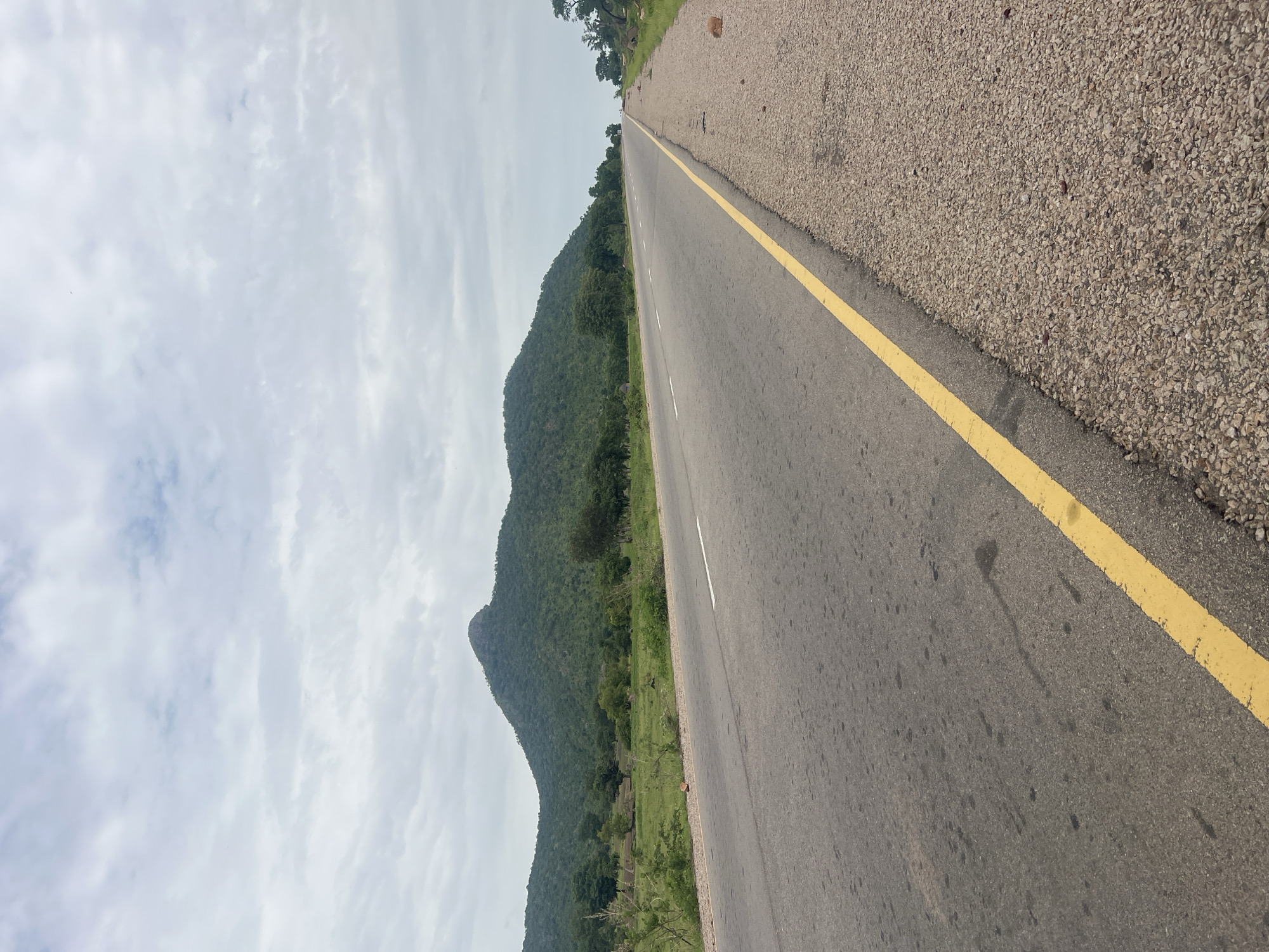The media file shows a road stretching into the distance, bordered by a green, mountainous landscape on the left and a gravel shoulder on the right. The road itself is asphalt with a bright yellow line painted down its center, separating it into two lanes. White lines mark the edges of the lanes. The sky is overcast with a soft, diffused light.
The scene appears to be a rural or semi-rural area in Nigeria, given the location context. The mountains are covered in dense vegetation, suggesting a tropical or subtropical climate. The presence of a paved road indicates a connection to a town or city.
There are no people or discernible objects like vehicles visible in the image. The focus is on the landscape and the road itself. The time of day is difficult to ascertain precisely due to the overcast conditions, but it appears to be daytime. The overall mood is one of solitude and natural beauty, with the road serving as a pathway through the scenic environment. The gravel shoulder shows some scattered small stones, and the asphalt has some dark stains. No visible text is present in the image.