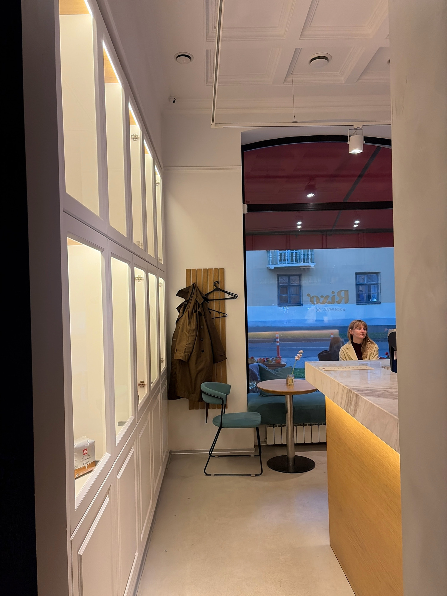 The image captures the interior of a modern coffee shop, likely located in Minsk, Belarus, as indicated by the presence of the "Rixa Espresso Bar" sign reflected in the exterior window. The foreground is dominated by a white marble counter with a warm wooden base illuminated by integrated lighting. To the left, a long wall features a series of illuminated display cabinets with white frames and glass fronts, some of which contain products, including a package of "Illy Classico" coffee. Further left, a dark, plain wall forms the edge of the frame.
In the mid-ground, a single teal chair with a black metal frame sits beside a small, round wooden table. A coat rack hangs on the wall behind the chair, displaying a long, brown trench coat. Through a large window, the exterior scene is visible, showing a street with parked cars and building facades. A young woman with blonde hair, wearing a light-colored jacket over a dark top, is seated at the counter, looking towards the right. She appears to be a customer or an employee of the establishment.
The ceiling is a patterned white structure with recessed lighting and circular fixtures, one of which appears to be a smoke detector. The overall ambiance is clean, contemporary, and well-lit, suggesting a welcoming space for patrons. The floor is a smooth, light-colored concrete or epoxy material.