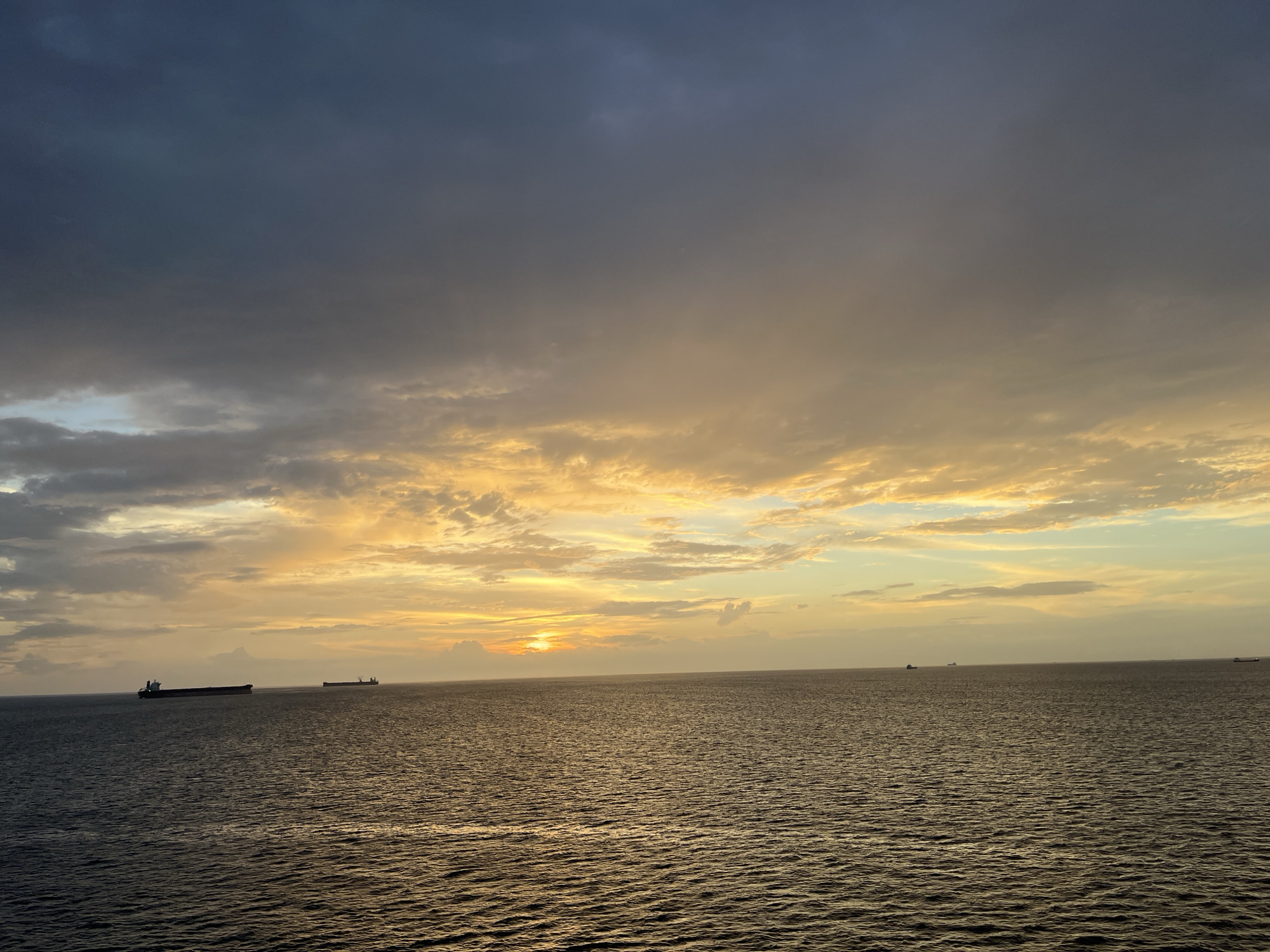 The image presents a serene oceanic landscape at either sunrise or sunset. The horizon divides the frame into two primary elements: a vast expanse of water in the lower half and a dramatic sky above.
The sea, a deep and dark hue, reflects the warm colors of the sky in its gentle ripples. Several large cargo or container ships are visible on the horizon, appearing as dark silhouettes against the bright light. Two are notably distinct: one on the far left and another slightly to its right, with additional smaller vessels discernible further in the distance.
The sky is a prominent feature, showcasing a remarkable gradient of colors. The upper portion is dominated by heavy, dark grey and bluish clouds. As the sky descends towards the horizon, these clouds thin and transform into brilliant shades of orange, yellow, and gold, illuminated by the low-lying sun. The sun itself is partially obscured by clouds on the horizon, casting an intense, warm glow that spreads across the mid-section of the sky. Breaks in the clouds allow rays of light to filter through, enhancing the dramatic effect.
The scene conveys a sense of calm and vastness on the open ocean. The time of day is clearly indicated by the intense golden and orange light, characteristic of dawn or dusk. No people or visible text are present in the image. The weather appears clear over the water, with significant cloud cover in the sky.