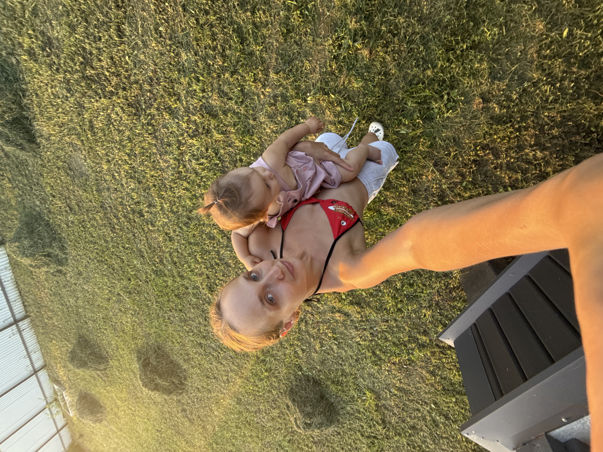 A woman and a baby are on a grassy lawn. The woman is wearing a red bikini top and grey shorts, and she is holding the baby, who is wearing a pink outfit. The background shows a green lawn with some haystacks and a structure with a corrugated roof. The scene appears to be in the village of Podshivalovo, Russia, during daylight hours.