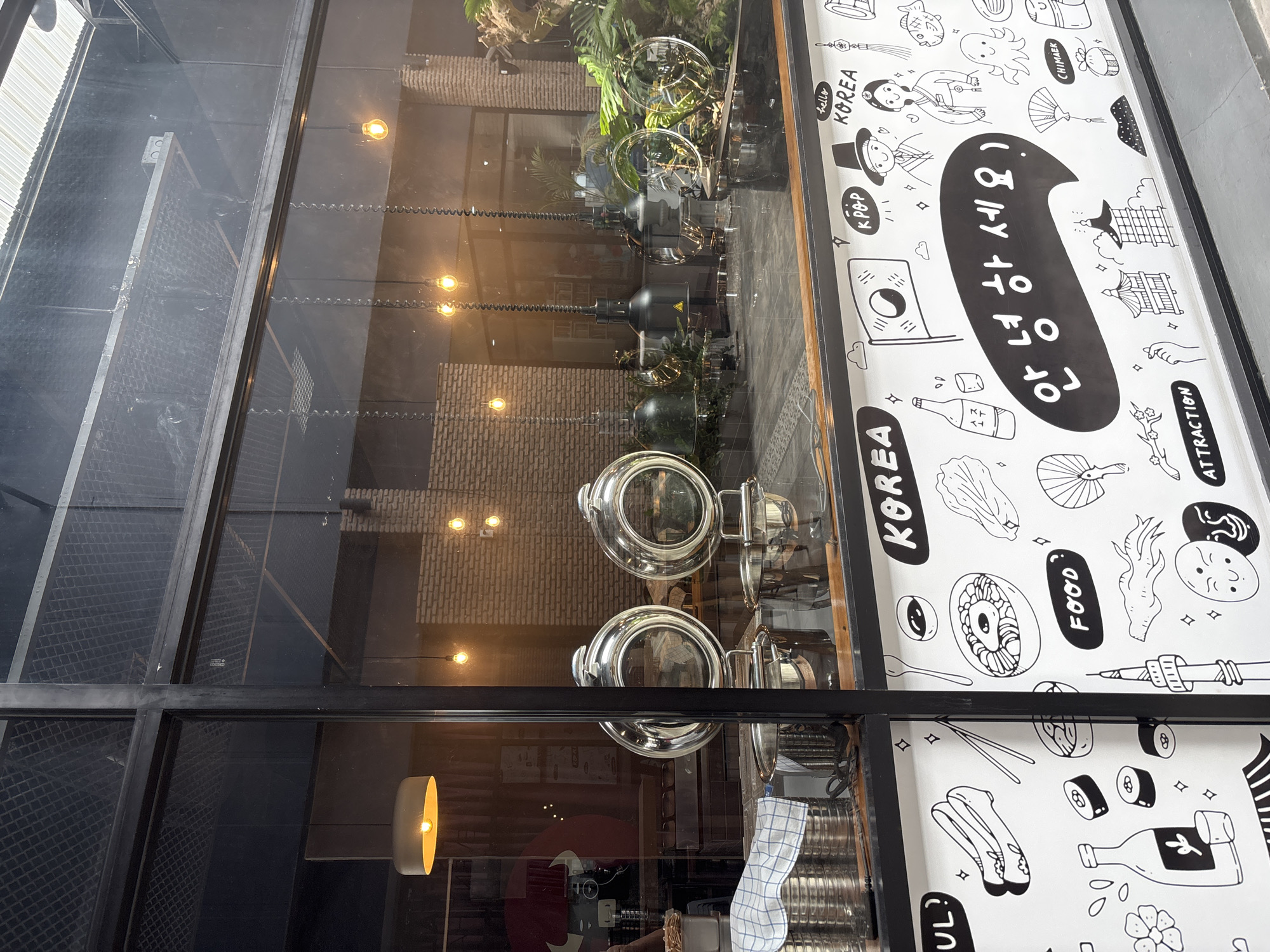 The image shows a window display, likely of a Korean restaurant. The window is divided into panes by a black metal frame. Through the glass, the interior of the restaurant is visible, featuring pendant lights, foliage, and dining setups. The right side of the window pane is covered in black and white Korean-themed doodles and text, including the Korean greeting "안녕하세요" (annyeonghaseyo), the words "KOREA," "FOOD," "ATTRACTION," "KPOP," and "CHIMAEK". The location seems to be Baan Chaweng Noi, Thailand.