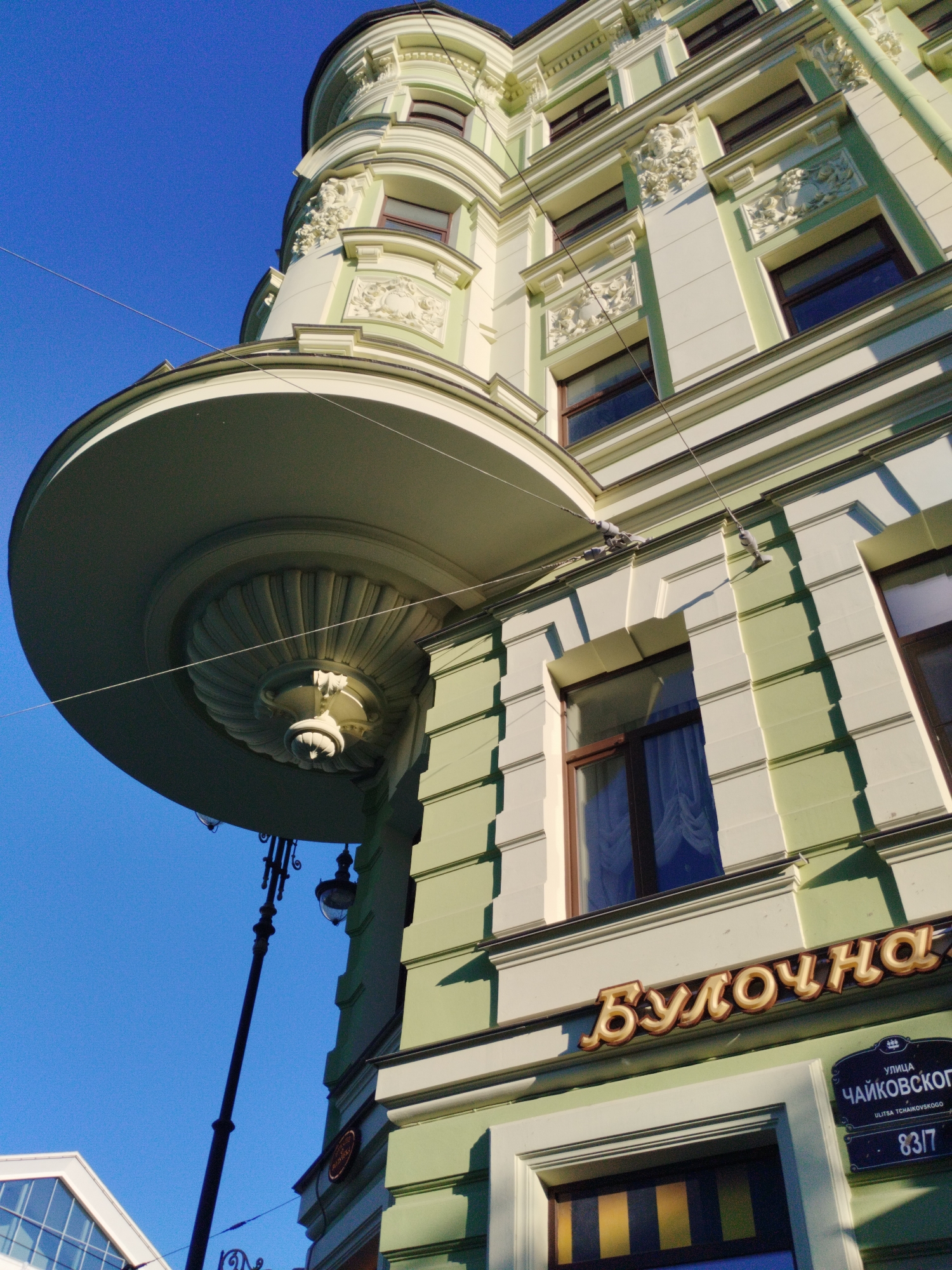 The image shows a low-angle view of the facade of an ornate, light green building in Saint Petersburg, Russia. The building features classical architectural details, including decorative relief sculptures and elaborate cornices. A prominent curved balcony, supported by a fluted column and decorative corbels, juts out from the upper story. The windows are framed with white molding, and some have blue curtains visible. A sign in Cyrillic script, reading "Булочная" (Bakery) in gold lettering, is mounted above a street sign that identifies the location as "УЛИЦА ЧАЙКОВСКОГО" (Tchaikovsky Street) and provides the address "83/7". A black lamppost with decorative elements stands to the left of the building, extending upwards into the clear blue sky. The lighting suggests it is daytime, possibly late afternoon, with strong sunlight casting shadows.