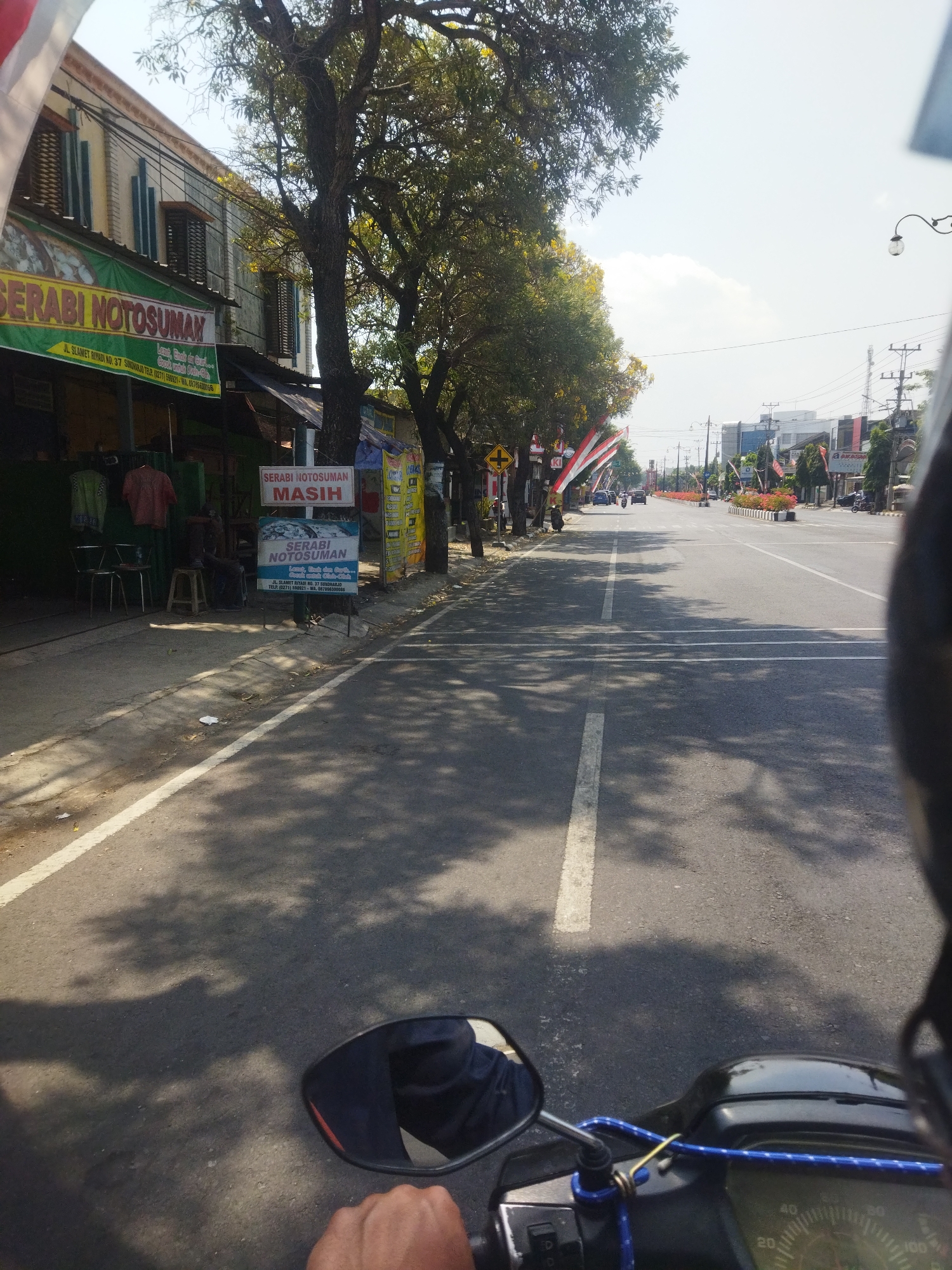 The image captures a street scene in Sukoharjo, Indonesia, as viewed from the perspective of someone riding a motorcycle. The road is paved with asphalt and marked with white lines, indicating a roadway with at least two lanes in each direction. Sunlight casts shadows from trees onto the road surface, suggesting a daytime setting.
On the left side of the road, there is a row of buildings with shop fronts. One prominent shop has a green awning with "SERABI NOTOSUMAN" written in large yellow letters. Below this, smaller text indicates the address "JL. SLAMET RIYADI NO. 37 SUKOHARJO" and a phone number. Several items of clothing are displayed on hangers outside this shop. Another sign for "SERABI NOTOSUMAN" is posted on a stand, with smaller text and an image of what appears to be food.
Further down the street, more shops and buildings are visible, with trees lining the roadside. Several Indonesian flags, red and white, are displayed along the street, possibly indicating a national holiday or event. Traffic is present, with a few cars and motorcycles visible on the road ahead.
On the right side of the image, a motorcycle's handlebar and a portion of the instrument panel are visible, along with a rider's hand, confirming the point of view. A blue elastic cord is attached to the handlebar. The rider is wearing a dark garment. In the background, more buildings and infrastructure, including a street light and utility poles, are present. The sky is bright and appears to be mostly clear with a few clouds.