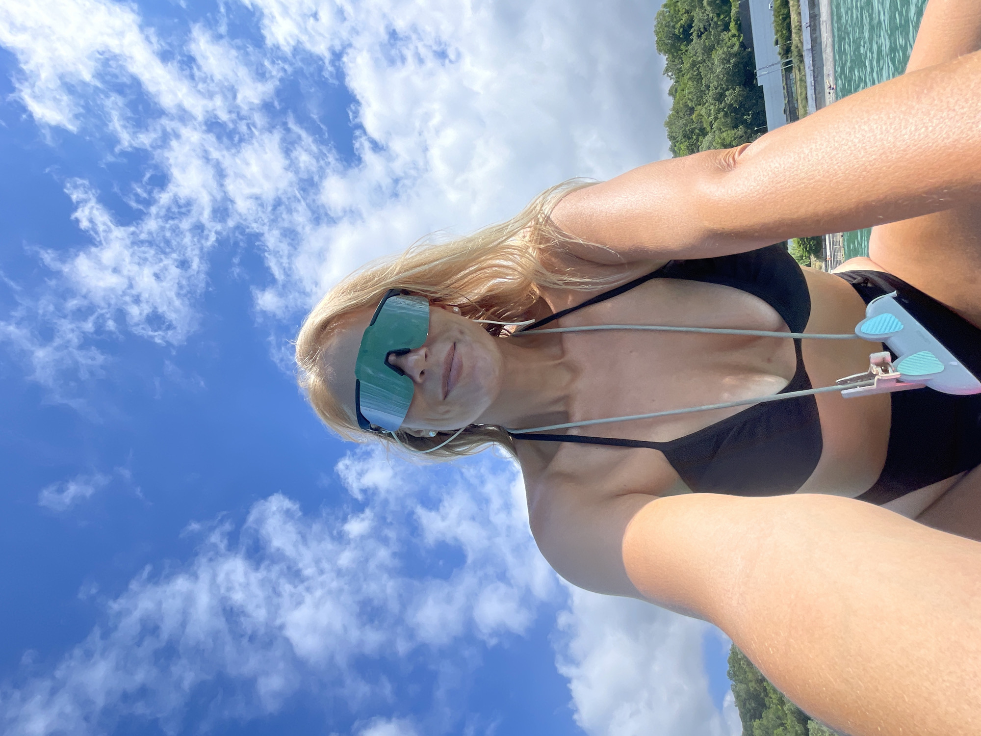 A woman with blonde hair, wearing a black bikini and large, futuristic green-tinted sunglasses, is taking a selfie outdoors. Her arms are crossed, and she is looking at the camera. The image is taken from a low angle, looking up at the sky, which is a vibrant blue with scattered white clouds. The woman's skin is tanned, suggesting a sunny day.
In the background, out of focus, is a body of water that appears to be a canal or harbor, with a concrete walkway and railings visible along its edge. Beyond that, lush green trees cover rolling hills, indicating a natural, possibly tropical or subtropical, environment. The water reflects the sky and trees. The overall impression is one of a warm, bright day, likely during daylight hours, with the woman enjoying a recreational activity near the water. The woman's expression is neutral, but the context suggests a relaxed and enjoyable atmosphere. There is no visible text in the image.