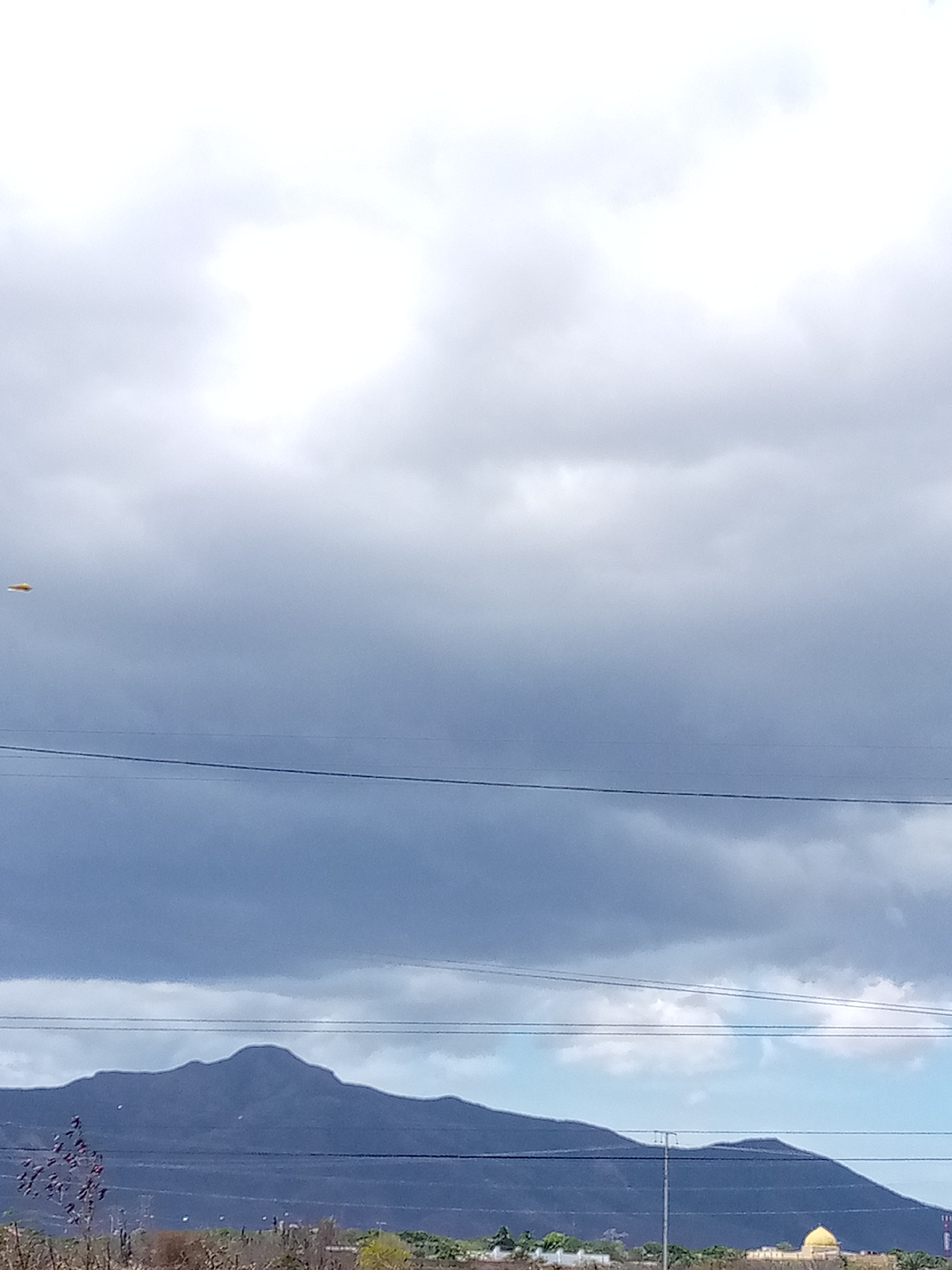 The image shows a cloudy sky above a landscape in Las Guevaras, Venezuela. A mountain range is visible in the middle distance, with power lines running across the frame. In the lower right, a building with a golden dome stands among sparse vegetation. A yellow, disc-shaped object appears in the upper left sky.
