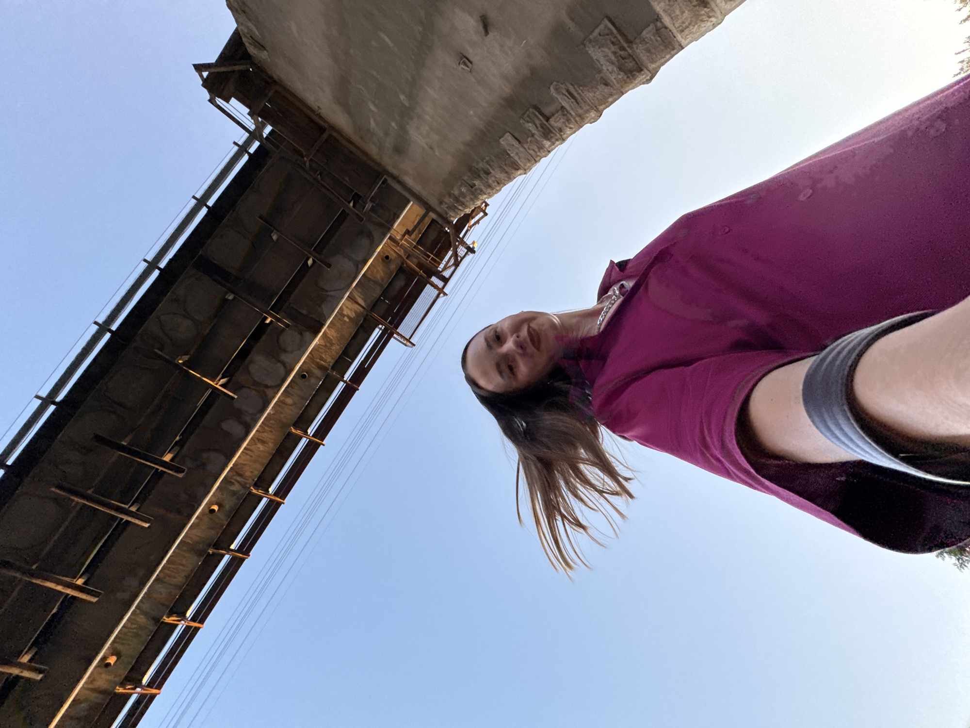 A low-angle, wide shot captures a person standing beneath a large, concrete structure that appears to be a bridge or overpass. The underside of the structure is visible, with its rough, textured concrete and the intricate metal supports and beams of the bridge's undercarriage. The structure extends diagonally across the left side of the frame, creating a stark geometric contrast with the clear, bright blue sky that occupies the rest of the image.
A young woman is the primary human subject, her face turned towards the camera in a selfie-style shot. She is wearing a long-sleeved, deep purple garment, possibly a jacket or a large shirt, and a grey band is visible around her waist, suggesting she might be wearing shorts or athletic wear. Her medium-length, brown hair is blowing outwards, adding a sense of movement and dynamism to the scene. A delicate silver chain necklace is visible around her neck. She appears relaxed and perhaps amused, her gaze directed at the camera.
The scene is lit by bright daylight, suggesting it is either late morning or early afternoon. The clear sky indicates fair weather. The overall impression is one of an urban or semi-urban environment, possibly near a transportation hub, given the presence of the large overpass. The low angle makes the viewer feel as if they are looking up from ground level, emphasizing the scale of the structure and the expanse of the sky. There is no visible text in the image.