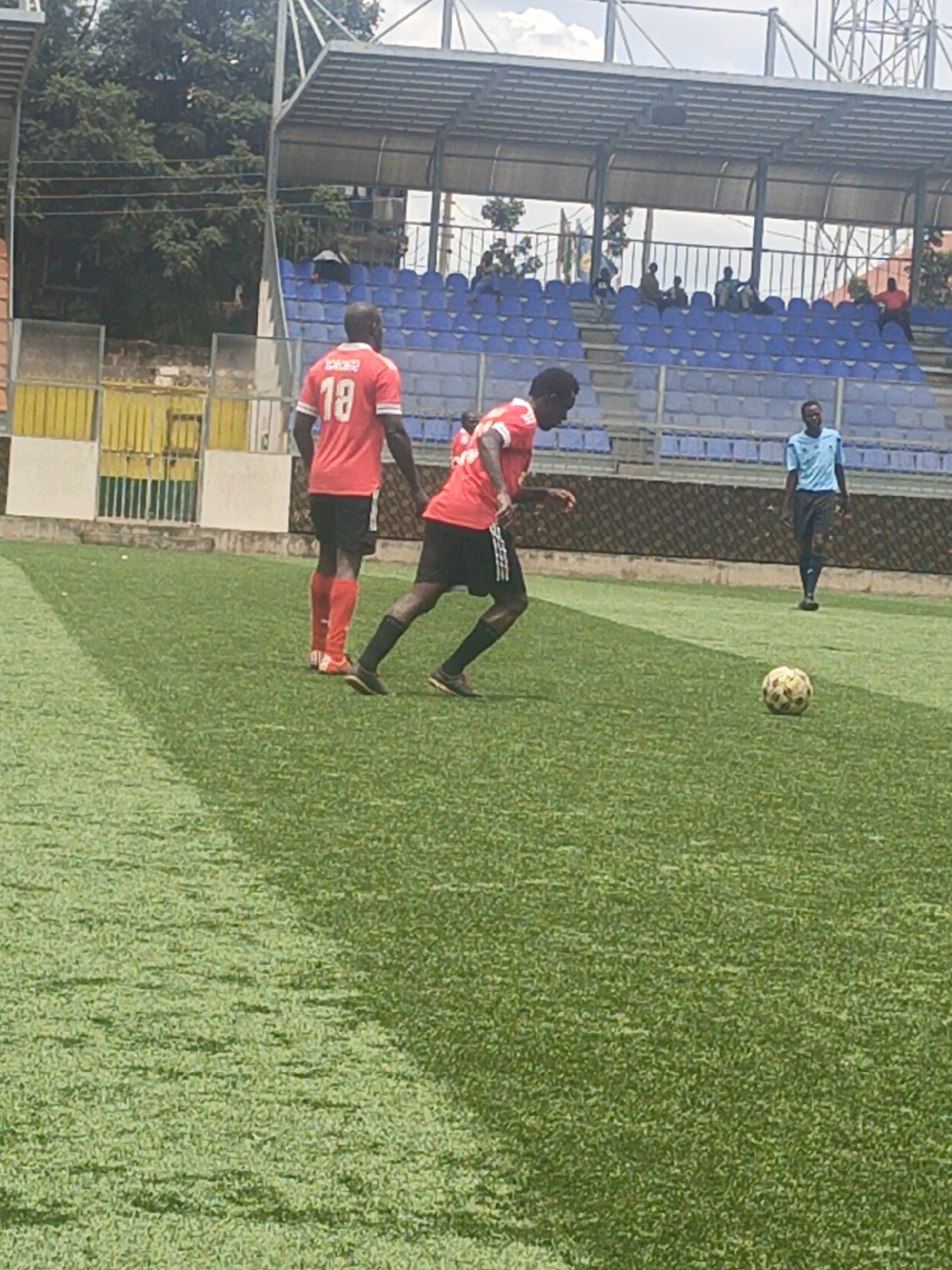 Two soccer players, one wearing a red jersey with the number 18 on the back, are on a green soccer field. One player is standing, and the other is running toward a soccer ball. A referee in a blue uniform is walking behind them. Behind the players is a stadium with blue seats under a metal roof. The stadium is in Nairobi, Kenya.