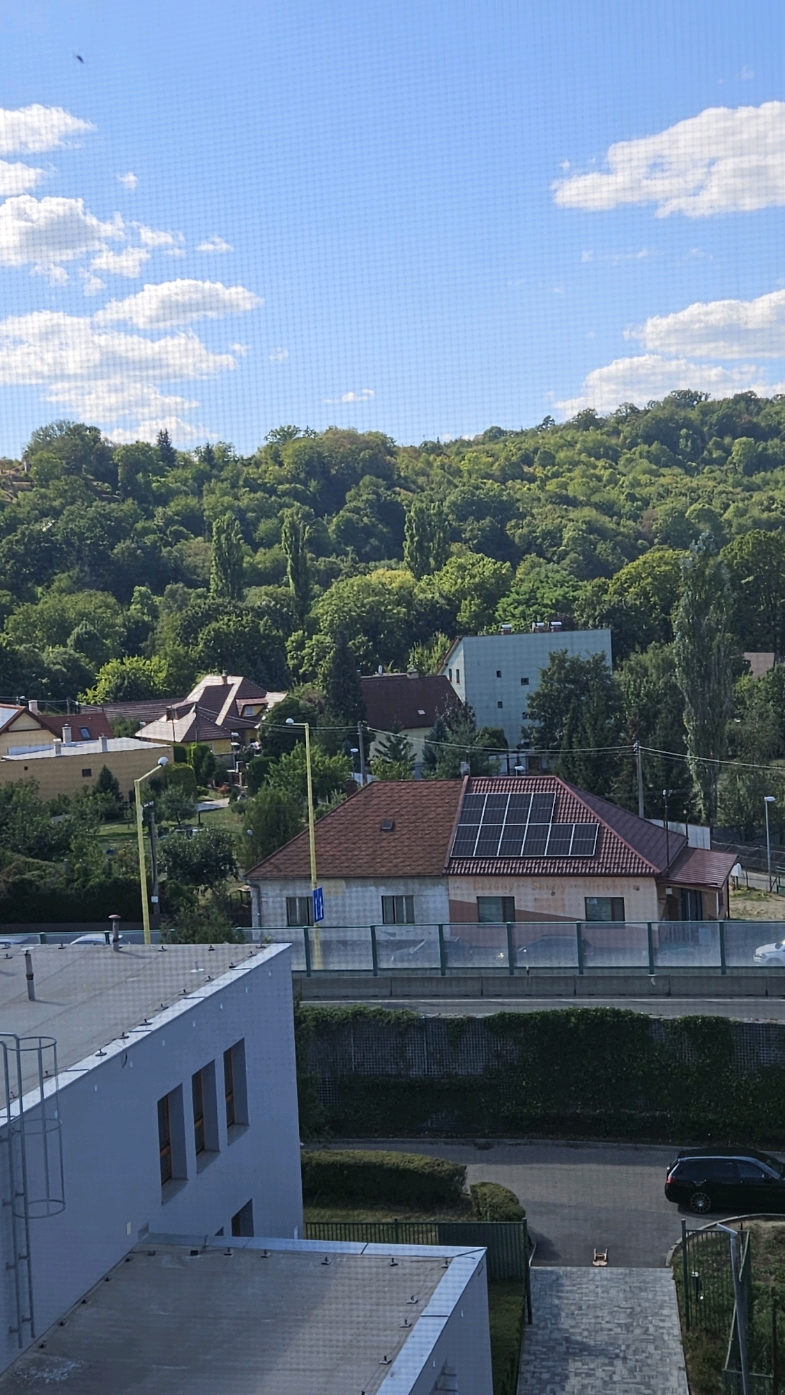 The image captures a view from an elevated position overlooking a residential area and a wooded hillside under a partly cloudy sky. In the foreground, a portion of a modern, light-colored building with multiple windows is visible. Adjacent to it is another building with a flat, dark-colored roof.
Further into the scene, a house with a reddish-brown roof featuring solar panels is prominent. Several other houses with red or yellow roofs are nestled among lush green trees. A light-colored building with a grid-like facade and rectangular windows is situated on the slope behind these houses.
The background is dominated by a densely wooded hillside. A street runs across the midground, bordered by a green hedge and a fence. A black car is parked on the right side of the street, and another car is visible in the distance on the left. A tall, yellow pole with a light fixture stands near the house with solar panels. The overall impression is that of a suburban or semi-rural environment.
While the image itself does not provide definitive landmarks to pinpoint a specific location within Prešov, Slovakia, the architectural styles of the houses and the vegetation are consistent with residential areas in the region. The presence of solar panels suggests a modern adaptation to housing in the area.