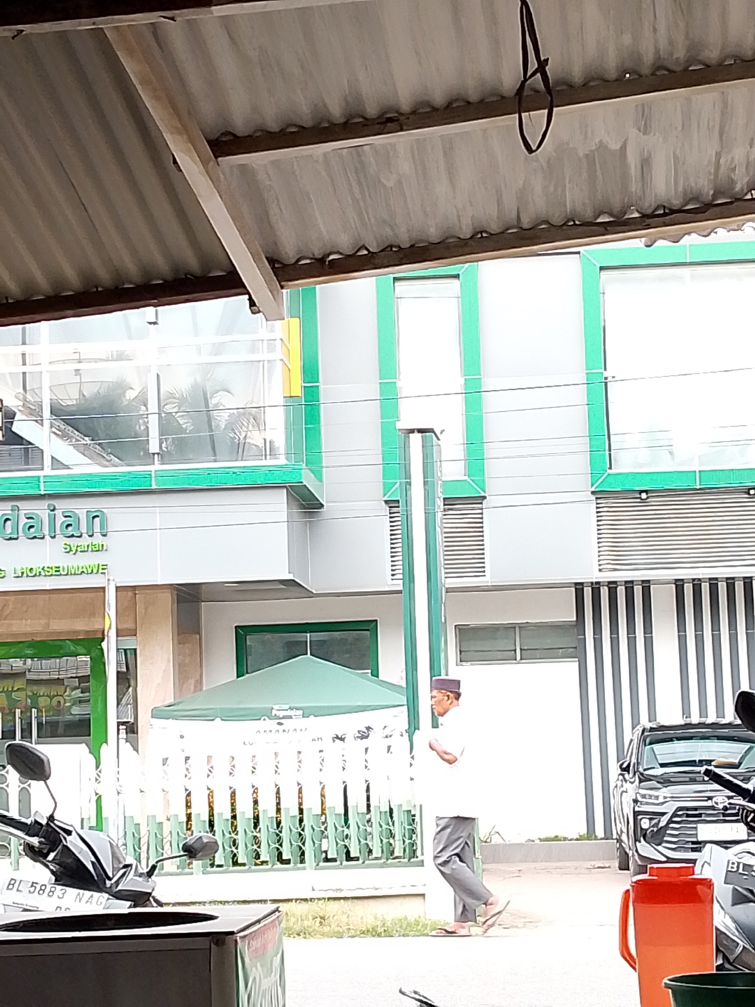 The image shows an outdoor scene in front of a modern building with green and white accents. In the foreground, a motorcycle is partially visible. A man wearing a white shirt, grey pants, and a dark cap is walking towards the right side of the frame. Behind him, a black SUV is parked. An orange jug is also visible in the foreground, near the motorcycle. The building has large windows and appears to be an office or commercial establishment. Some text is visible on the building, including "daian Syariah" and "G LHOKSEUMAWE". The scene suggests a daytime setting, possibly in Lhokseumawe, Indonesia, as indicated by the text. The overall impression is that of a daily activity in a commercial or institutional area.