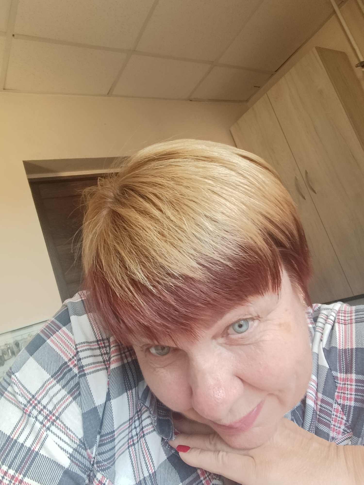 This is a close-up, slightly angled selfie of a middle-aged Caucasian woman with short, layered blonde hair with reddish undertones. She has light blue eyes and is wearing a plaid shirt with white, grey, and red patterns. Her left hand is resting on her chin, and her fingernails are painted red. The background appears to be an indoor environment, possibly an office or room, with a suspended ceiling with square panels and a wooden cabinet with two doors visible on the right. The lighting suggests an interior setting, and there's no clear indication of the specific location within Svietlahorsk, Belarus, beyond it being indoors.