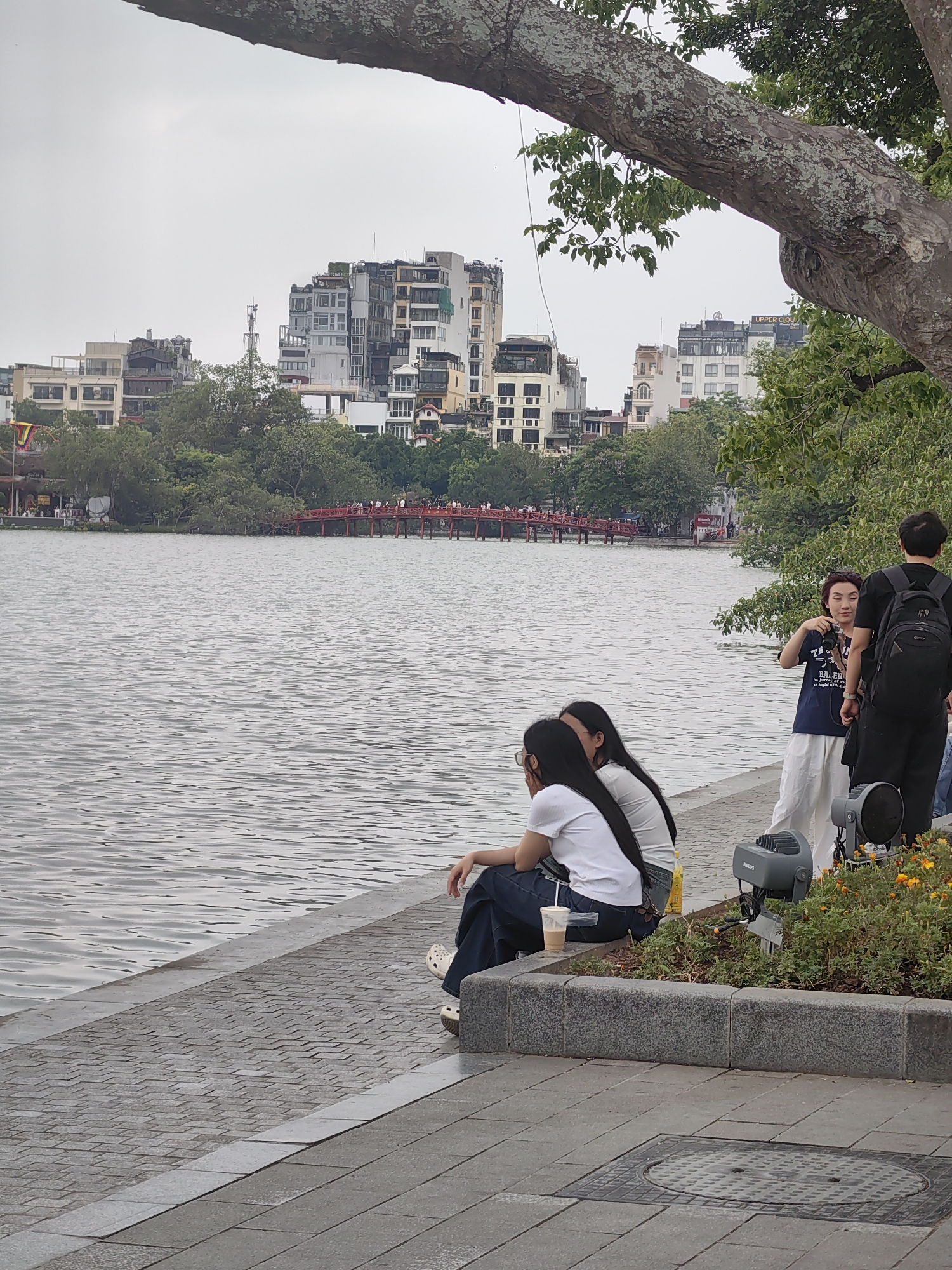 The image presents a daytime scene by a lake in an urban environment, likely Hoan Kiem Lake in Hanoi, Vietnam, under an overcast sky.
In the foreground, a substantial tree branch stretches diagonally across the top of the frame. Below it, a paved walkway lines the lake's edge. Two young women with long dark hair sit on the edge of the walkway, facing the water, with a plastic cup containing a drink next to one of them. To their right, a woman wearing a dark blue t-shirt with the visible text "BA LEN" and partially legible words below, stands holding a camera, seemingly photographing the lake. Next to her, a man with a black backpack stands with his back to the viewer. In a flowerbed beside the walkway, two grey Philips spotlights are visible.
The midground features the calm, rippled surface of the lake. Across the water, the distinctive red wooden Huc Bridge is visible in the distance, with several people walking across it. Beyond the bridge and surrounding foliage, the background showcases a dense cityscape composed of various multi-story buildings. On one of these buildings, part of a sign reads "UPPER CLOU".
The overall scene depicts people relaxing by the lakeside, observing the view, and engaging in photography, set against a backdrop of a bustling city.
