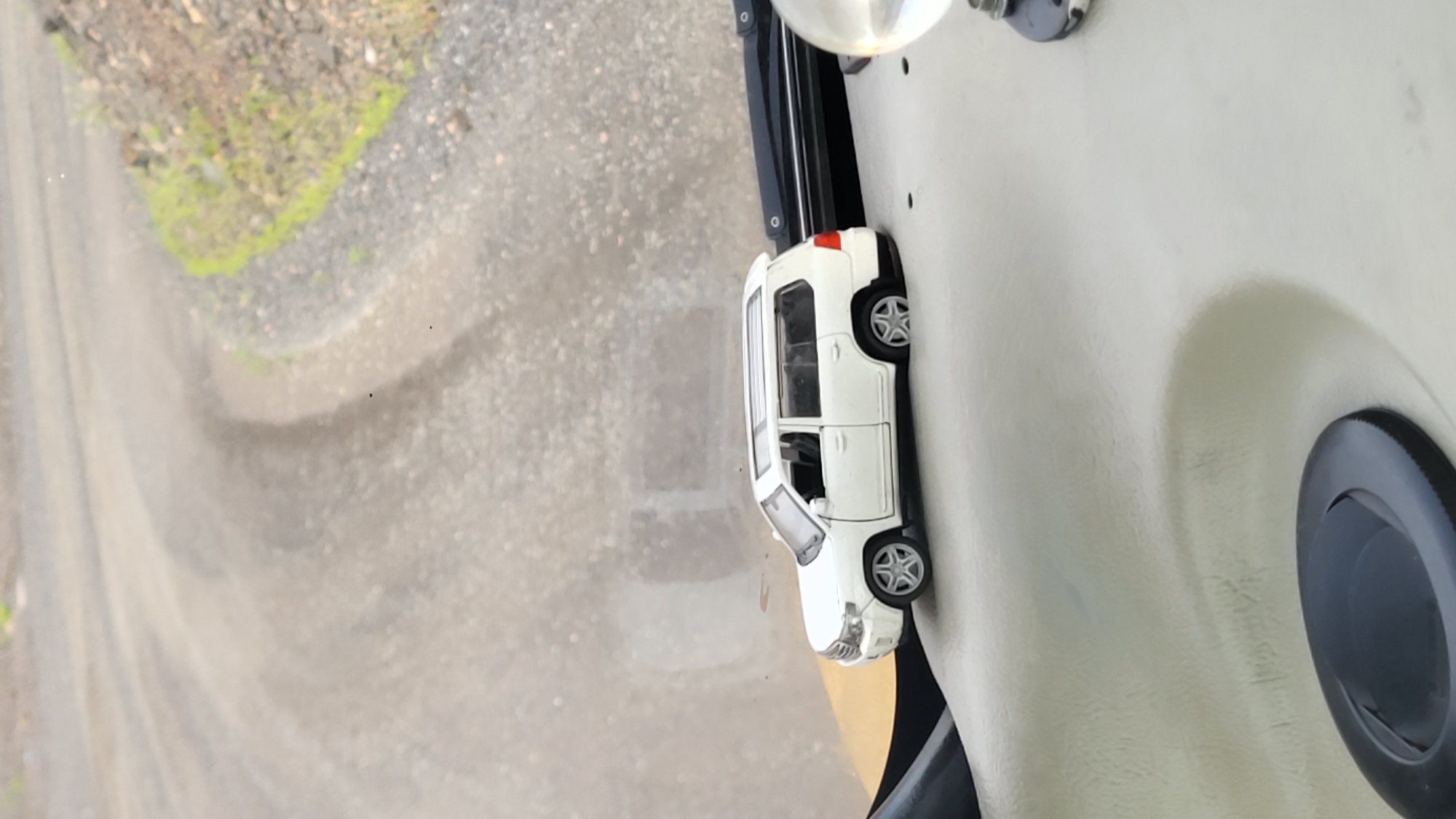 A miniature white SUV toy car is positioned on a textured, light-colored surface, possibly the dashboard of a vehicle. The car is facing left, with its wheels slightly turned. In the background, a blurred, dusty road curves to the right, bordered by rough terrain with scattered rocks and sparse green vegetation. The lighting suggests an outdoor, daytime setting. The specific location in Yerystivka, Ukraine, cannot be determined from the visual information provided.