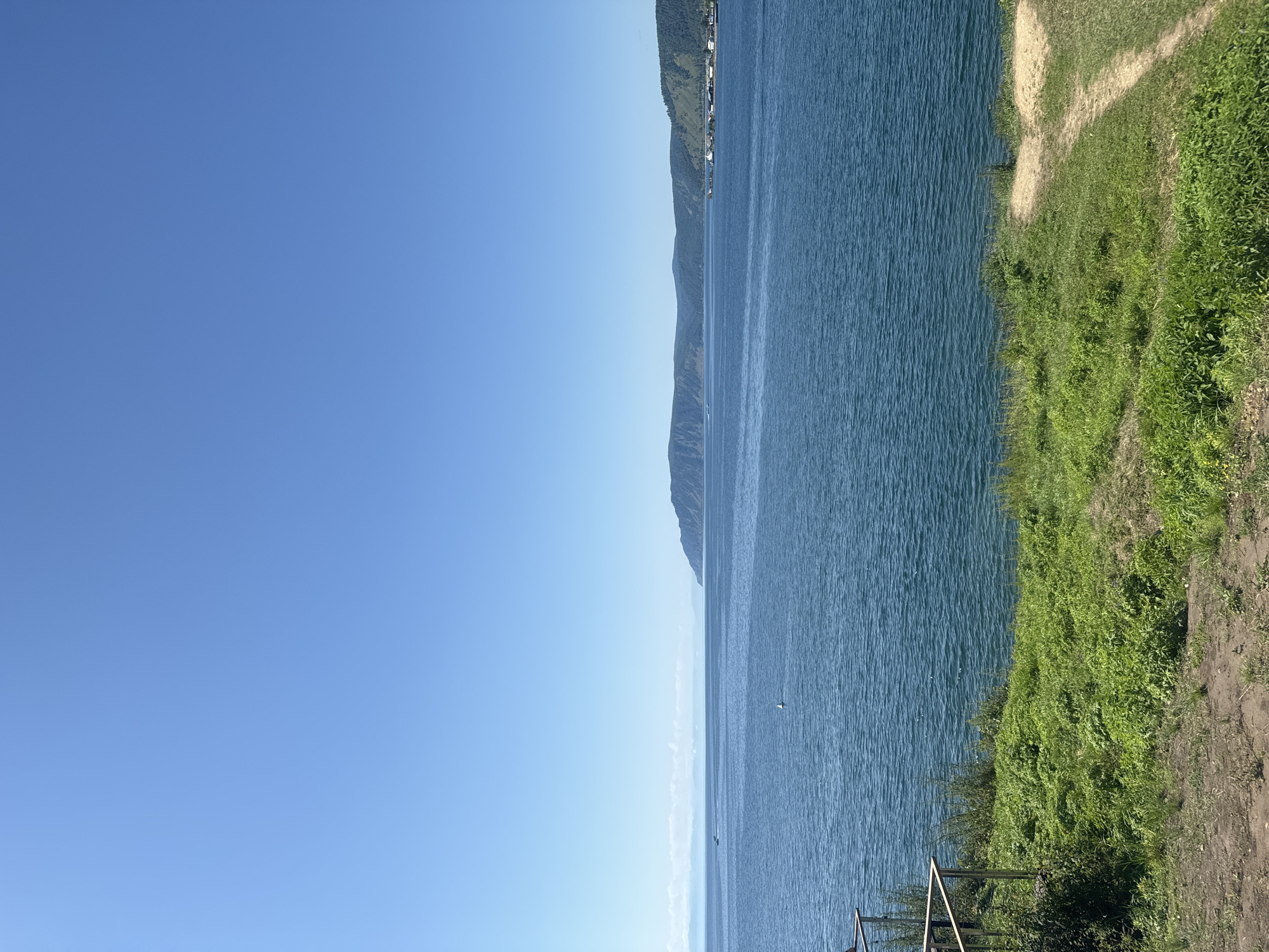 The image depicts a tranquil scene of Lake Baikal near Listvyanka, Russia, on a clear, sunny day. The foreground is dominated by a vibrant green, grassy embankment with patches of bare earth and a dirt path leading to the right. A metal railing runs along the left edge of the embankment. The expansive, blue waters of Lake Baikal stretch across the midground, with gentle ripples on its surface. In the distance, a chain of undulating, tree-covered hills forms the horizon, with a small settlement visible on the right side of the frame. The sky is a vast expanse of clear, bright blue, with no clouds in sight. There are no people or animals visible in the image.
