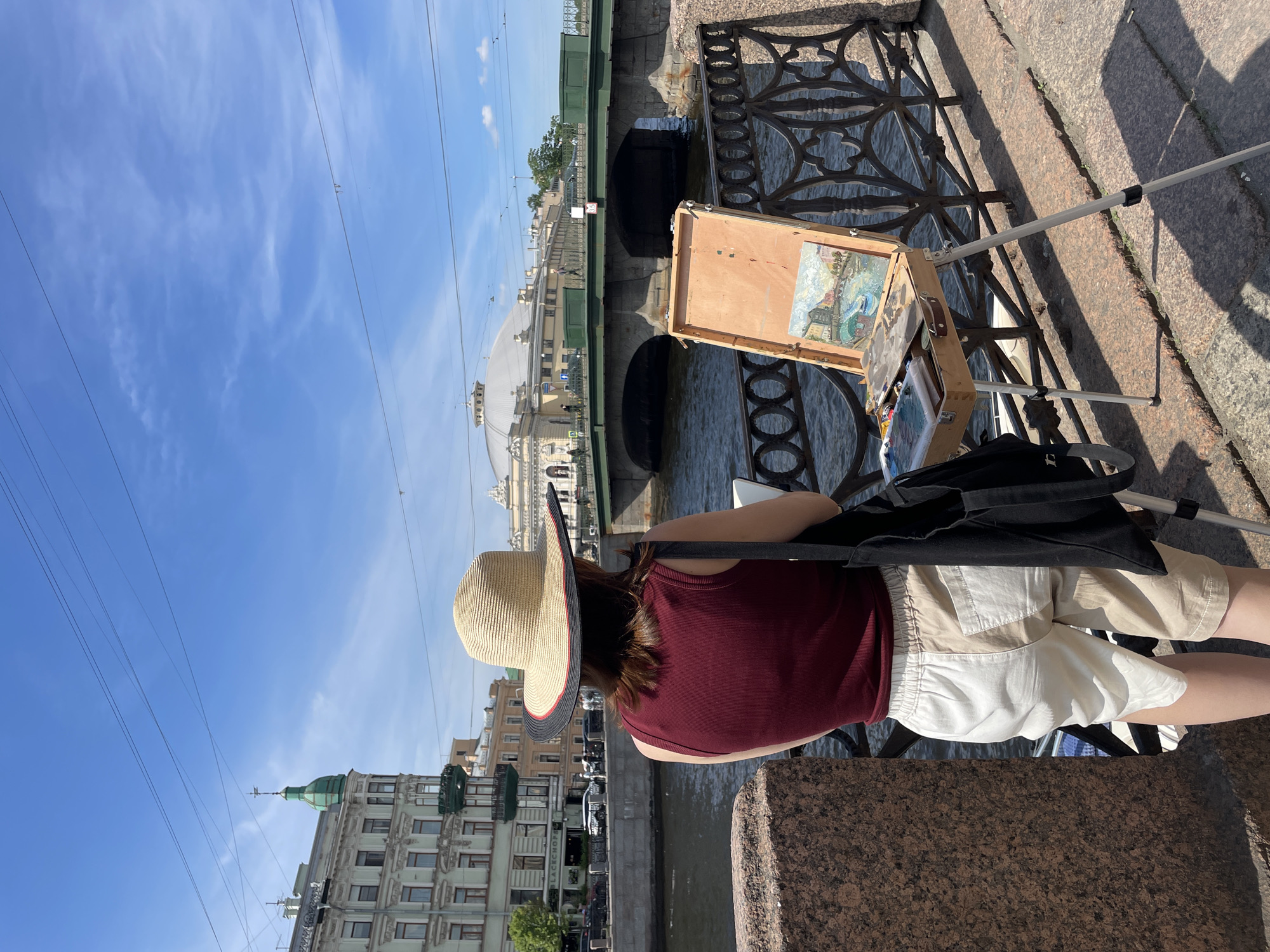 A person wearing a wide-brimmed straw hat and a maroon tank top is standing on a stone embankment, facing away from the camera. They are also wearing beige and white shorts. In front of them, an easel holds a small painting of a cityscape, likely a canal scene with buildings. A black bag rests on the ground behind the easel. The background shows a canal with water, a green bridge spanning it, and buildings on the far side. In the distance, a large domed building is visible, characteristic of Saint Petersburg architecture. The sky is blue with wispy white clouds, and power lines are strung across the top of the frame. The lighting suggests it is daytime, with strong shadows cast by the sun. The scene appears to be outdoors in an urban environment, likely near a body of water, consistent with the canals of Saint Petersburg. The person seems engaged in painting or observing their artwork, suggesting an artistic activity. There is a sign on the bridge with Cyrillic text, and a sign on the building to the left with "BLACKSHORE" visible.