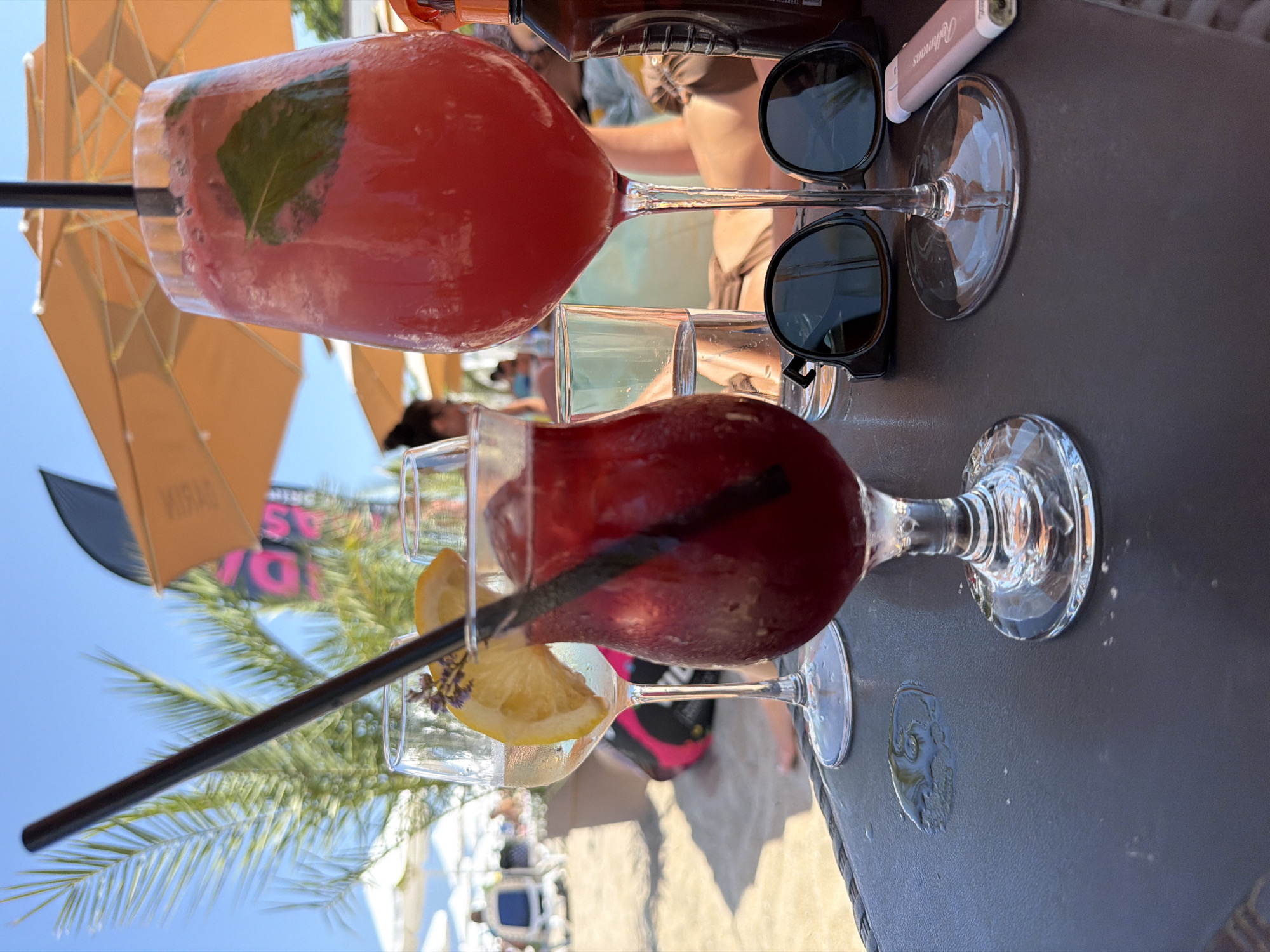 The image depicts a close-up view of a table at a beachside establishment in Sozopol, Bulgaria, on a sunny day. The table is dark and textured, with a few water spots visible. In the foreground, three drinks are present. On the left, a glass of red liquid with ice and a black straw, garnished with a slice of lemon and a sprig of lavender, is prominently displayed. To its right, a large wine glass holds a vibrant red beverage with ice and a green leaf, also with a black straw. Behind these, a clear glass with water is visible.
On the table, a pair of dark sunglasses rests near the base of the wine glass. To the right of the sunglasses, a pink lighter is placed. In the background, palm fronds and an orange umbrella suggest a tropical or beach setting. Other beachgoers are faintly visible in the distance, some seated and others walking, indicating a public beach environment. A bottle of sunscreen is also partially visible on the right side of the image. The overall scene conveys a relaxed, summery atmosphere at a beach bar in Sozopol.