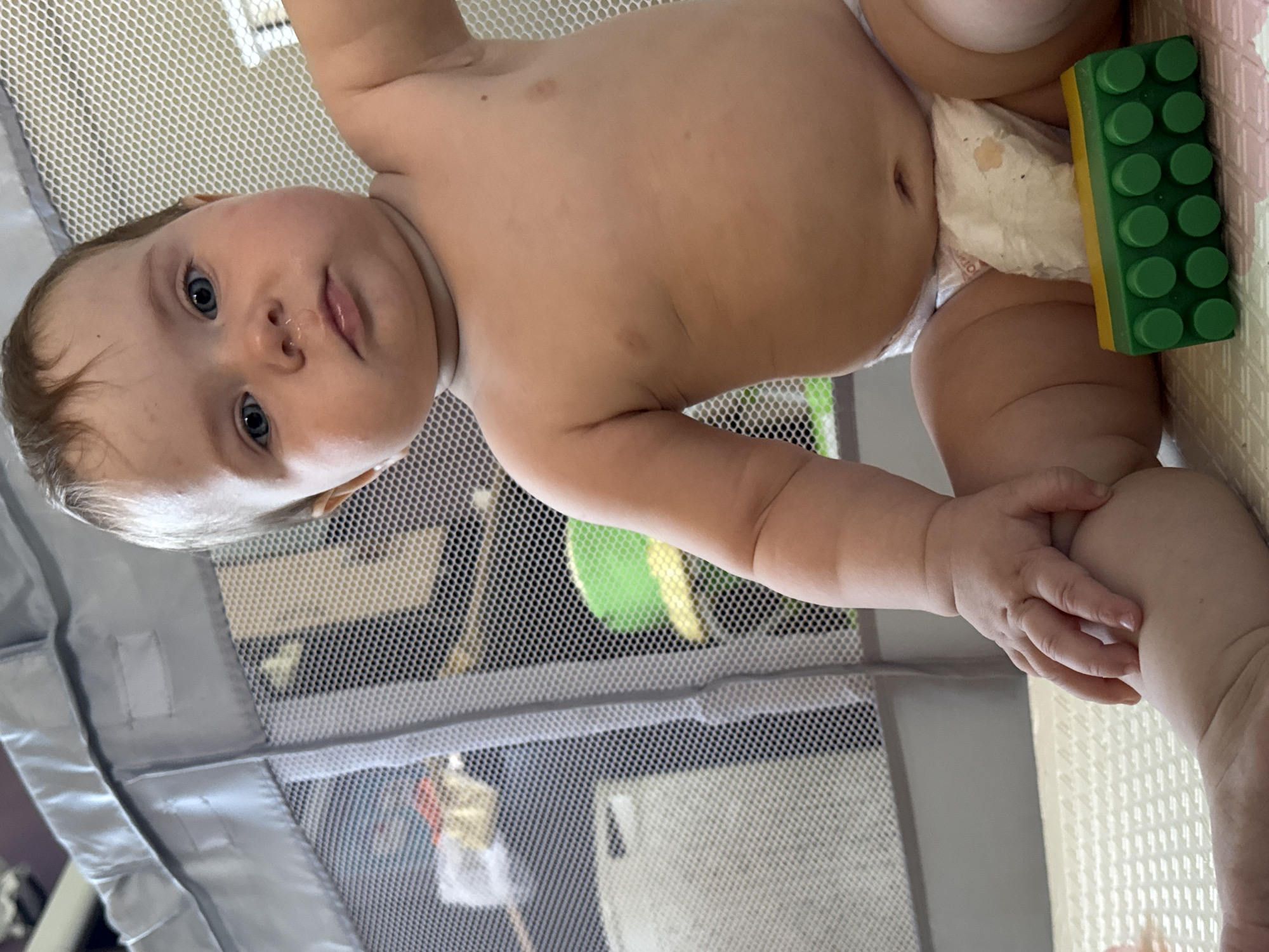 A baby wearing a diaper is lying on a playmat, with a green and yellow Lego block near the diaper. The baby is in a playpen. The location is not identifiable.