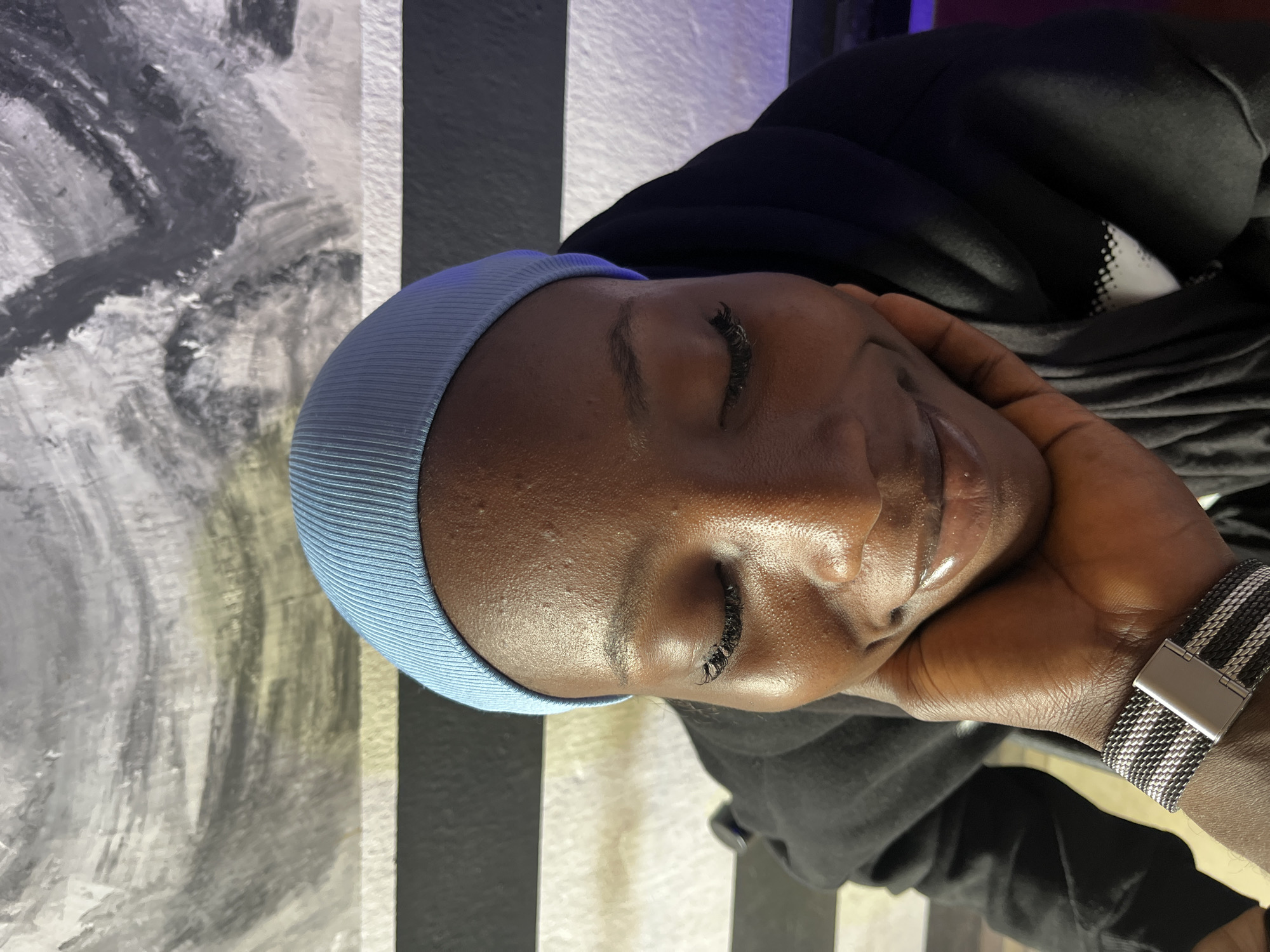 The image captures a close-up portrait of a person with dark skin, seemingly in a state of rest or contemplation, taken in Jos, Nigeria. The individual's eyes are closed, and their head is gently supported by their right hand.
The subject is wearing a light blue, ribbed cap or headwear, covering their hair, and a black hooded top or jacket. On their left wrist, a watch with a distinctive black and white striped mesh band and a silver clasp is visible. The lighting subtly highlights the natural sheen of their skin.
The background is composed of two different wall sections. To the left, an abstract artistic wall or large canvas features broad grey and white brushstrokes. To the right, a wall with prominent vertical black and white stripes can be seen. The overall setting appears to be an indoor space with decorative elements, suggesting a modern or stylized interior. There is no visible text in the image.