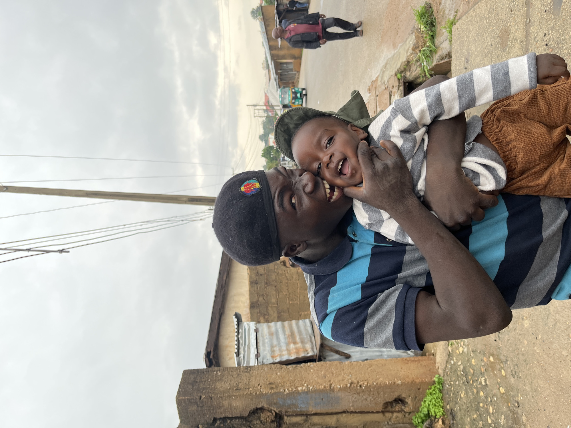 In this close-up, low-angle shot, two individuals, an adult and a child, are the central focus. The adult, wearing a black beret with a circular emblem and a blue and gray striped shirt, is holding the child. The child, dressed in a striped sweater, looks directly at the camera with a wide, joyous smile and open mouth, their face tilted upwards and towards the adult. The adult's hand is gently cupping the child's cheek, and they are also smiling. The scene appears to be outdoors, with a cloudy sky overhead. In the background, a street scene is visible, with a few buildings and what looks like a vehicle. The presence of a utility pole and wires suggests an urban or semi-urban environment. The overall mood is one of happiness and affection between the two subjects. The time of day appears to be overcast, possibly late afternoon. The location is identified as Jos, Nigeria.