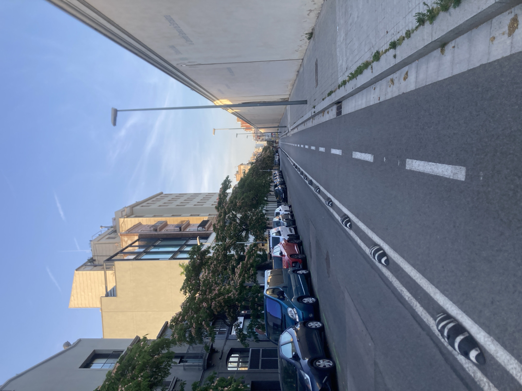 This is a street-level, medium-shot photograph taken in Barcelona, Spain, looking down a street with buildings on the left and a large, light-colored wall on the right. The sky is a clear, pale blue with faint wispy clouds. The street is paved and has a dedicated lane marked with white dashed lines, presumably for bicycles, running parallel to the parked cars on the left. Along the edge of this lane, there are several black and white striped speed bumps.
On the left side of the street, a row of parked cars is visible, including a dark blue hatchback, a teal van, a red SUV, a white van, and several other vehicles. The buildings on this side are multi-story, with a mix of architectural styles. Some have balconies, windows, and trees with pinkish-white blossoms are planted along the sidewalk, casting shadows on the street.
The right side of the street is dominated by a long, tall, light beige wall that extends into the distance. There are faint markings or text on the wall, possibly indicating a business or institution. A sidewalk runs alongside the wall, paved with rectangular concrete slabs and bordered by a concrete curb. Some sparse green vegetation is growing along the edge of the sidewalk. A single street lamp with a rectangular head is visible above the wall, on the right side of the frame.
The lighting suggests it is either early morning or late afternoon, with the sun casting long shadows and illuminating the buildings on the left. The overall scene depicts an urban environment with a focus on street infrastructure and a quiet, orderly atmosphere, with no people or active events captured.