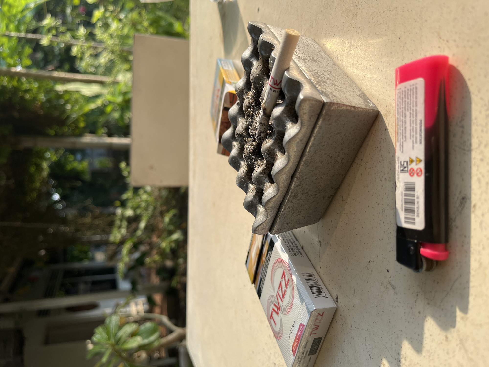 On a textured, light-colored outdoor table, a metal ashtray filled with ash and a single cigarette rests on its right side. To the left of the ashtray, a pack of "TWIZZ PRIME" cigarettes is visible, with the brand name prominently displayed in red lettering. A partially visible cigarette pack with yellow and orange detailing is positioned behind the ashtray. In the foreground, a black and pink disposable lighter lies flat on the table. The background shows a blurred outdoor scene with greenery and a building. The image appears to have been taken during daylight hours.