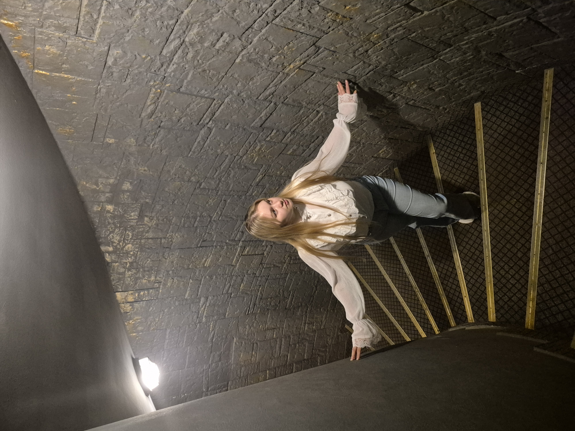 A young woman with blonde hair, wearing a white ruffled blouse and light blue jeans, stands on a spiral staircase. The staircase has dark grey carpet with a diamond pattern and yellow metal edging on each step. The walls of the staircase are textured to resemble dark stone blocks and are lit by an overhead light fixture. The woman is positioned on the middle section of the staircase, looking to her right with her arms outstretched, her hands lightly touching the wall and the railing. Her pose suggests she is either descending or ascending the stairs, or perhaps paused in transit. The location is identified as Chaikovsky, Russia.