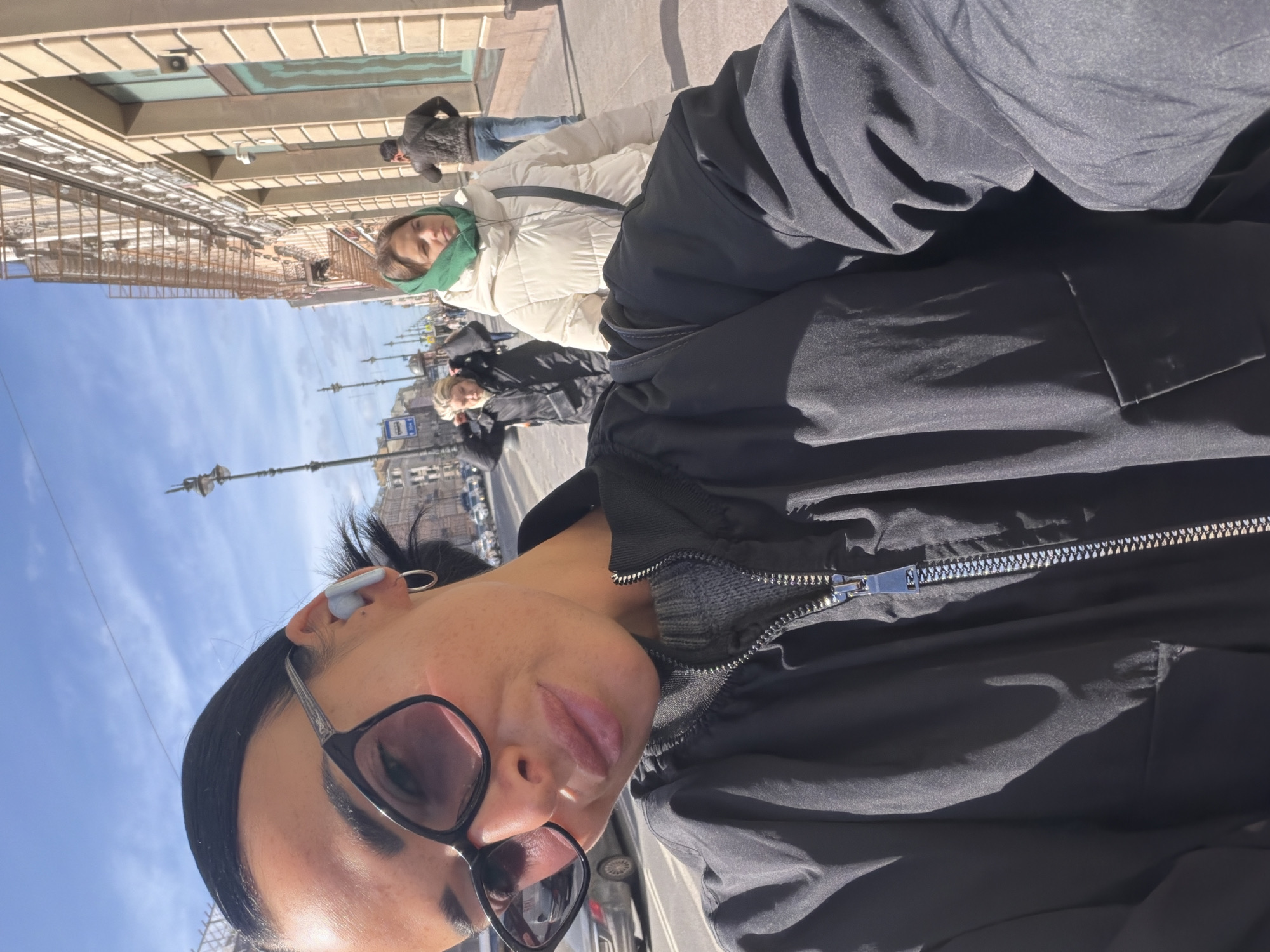 A selfie image was taken on a sunny day in Saint Petersburg, Russia. The picture includes a woman wearing sunglasses and an earpiece. She is wearing a black jacket. Behind her are other people walking on the sidewalk. There are street lamps. In the background, there are buildings and a clear blue sky.