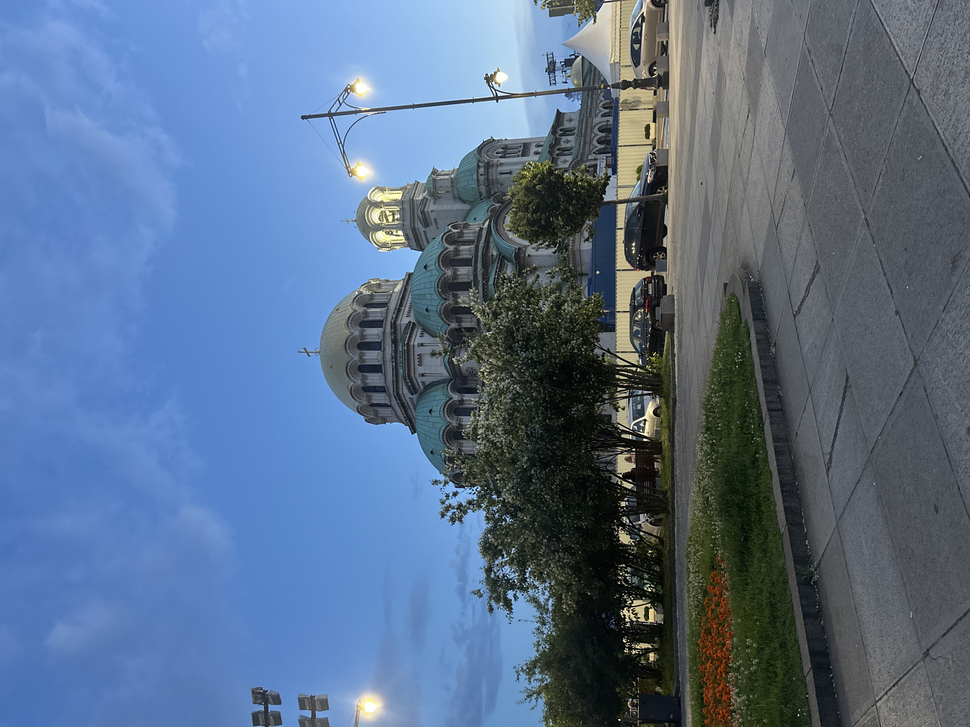 The image shows a large, ornate cathedral with multiple domes and spires under a twilight sky. The cathedral is light-colored with green domed roofs and intricate architectural details. In the foreground, there is a paved area with a low wall separating it from a grassy area with orange and white flowers. Several trees, some with white blossoms, are present in front of the cathedral. A street lamp with multiple lights illuminates the scene. Several cars are parked along the street, and two people are seated on a bench behind the trees. The overall scene suggests an outdoor public space, possibly a plaza or park, adjacent to a significant religious building, in the city of Unknown, Unknown.