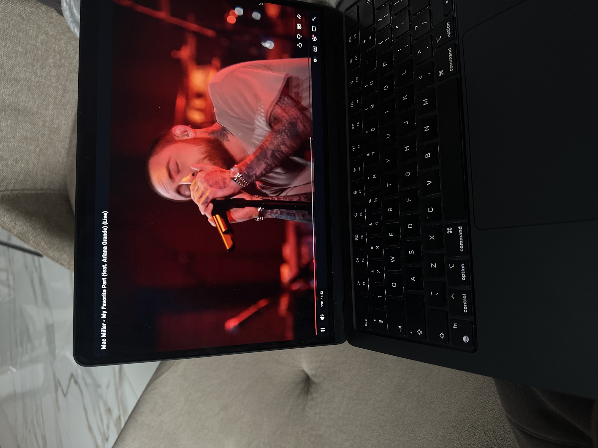 The image shows a laptop with a screen displaying a live performance of Mac Miller's "My Favorite Part" (featuring Ariana Grande). The laptop is black with a visible keyboard. The environment surrounding the laptop appears to be a neutral-colored upholstered surface. There is no indication of the image's location as Sofia, Bulgaria, or any other geographical information.