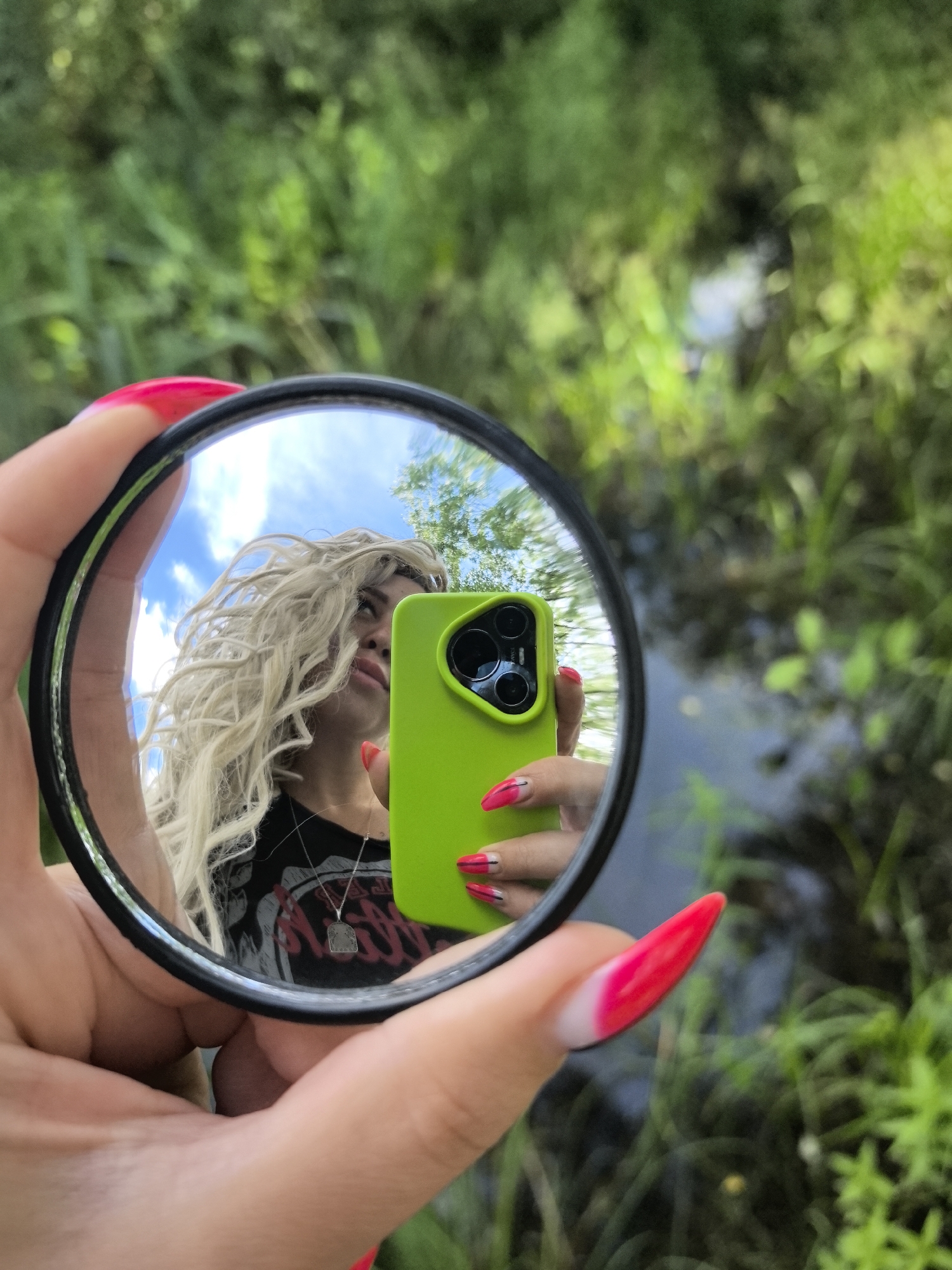 A person is holding a round mirror in their hand, with a bright pink and white-tipped fingernail visible. The mirror reflects the person's face, which has blonde, curly hair and is looking down. They are wearing a black t-shirt with a graphic design and a silver necklace. They are also holding a bright green smartphone. The background of the image is blurred green foliage and a body of water. The scene appears to be outdoors, likely in a natural setting. There is no information about the location in the city of Noviy, Russia.
