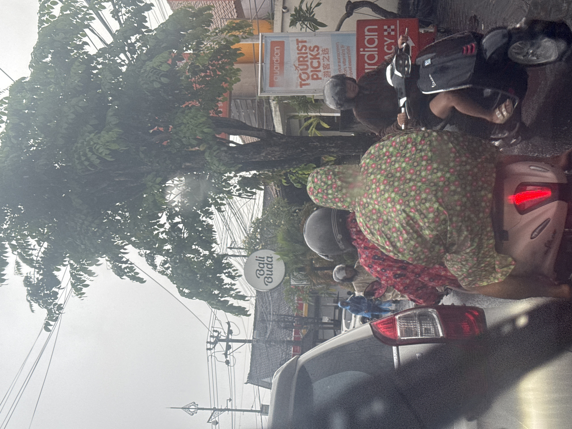 The image captures a bustling street scene in North Kuta, Indonesia, under an overcast sky, suggesting recent rain or impending showers. In the immediate foreground, the red taillight and bumper of a car are visible, indicating the perspective is from inside another vehicle or just behind it. Ahead, several individuals are stopped on motorbikes, characteristic of urban traffic in the region. One person, prominently featured, wears a green poncho with pink and white polka dots over a red patterned garment, also likely a rain cover, indicating the wet weather. Another person on a motorbike wears a grey helmet and a dark jacket, while further ahead, someone else is seen in blue rain gear.
Lush green trees with dense foliage line the street, with numerous power lines crisscrossing the upper portion of the frame against the cloudy sky. Buildings with tiled roofs are visible behind the trees and vehicles. Several commercial signs are mounted along the street. A prominent circular white sign displays "Bali Buda". Further down, a rectangular white sign with an orange "guardian" logo announces "TOURIST PICKS" and its Chinese translation "游客之选", with smaller text listing "your sunscreen on, vitamins & travel essentials". Another red sign, partially visible to the right, reads "rdian ACY", likely part of "Guardian Pharmacy". The scene conveys a sense of everyday urban life and traffic in Southeast Asia, with people adapted to the local weather conditions.