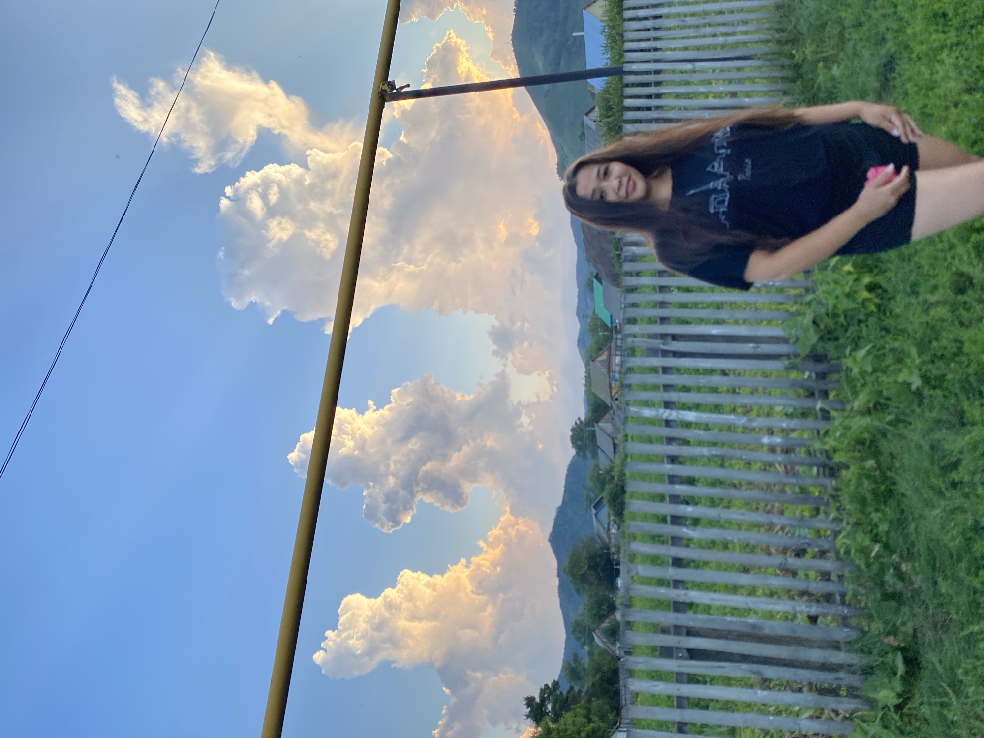 A young woman with long brown hair stands in front of a wooden fence in Maloarslangulovo, Russia. She is wearing a black t-shirt and black shorts. Behind her, houses and trees are visible, and in the distance, mountains rise under a sky filled with dramatic, illuminated clouds. A yellow metal pole runs horizontally across the upper portion of the image.