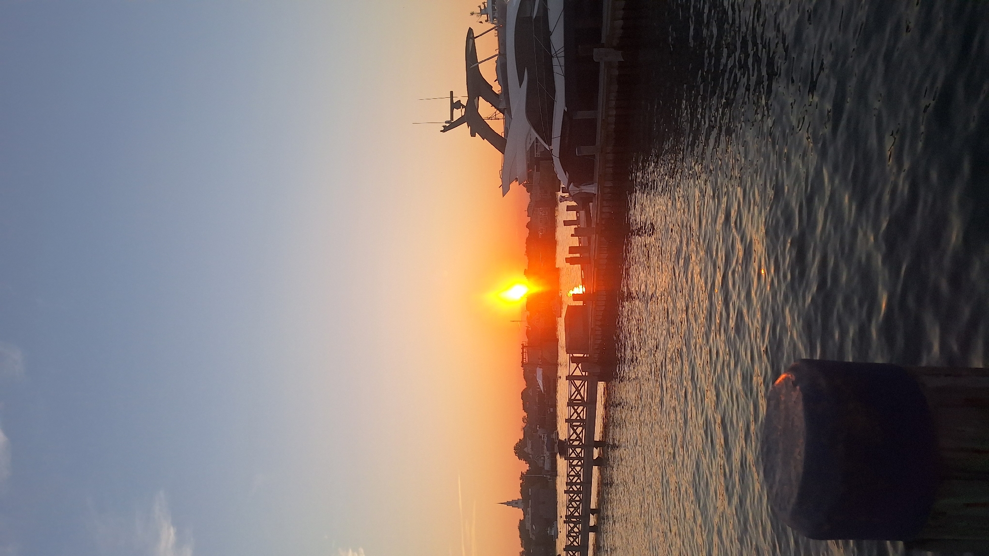 The image shows a sunset over Nantucket Harbor in Nantucket, Massachusetts, USA. The sun is low on the horizon, casting a warm orange glow across the sky and water. Several boats are docked in the harbor, including a large white yacht on the right side of the image. A wooden pier extends into the water in the foreground. In the background, the skyline of Nantucket town is visible, with a church steeple prominent among the buildings. The water is calm, with gentle ripples reflecting the colors of the sunset. The sky is a clear blue, fading into orange and pink near the horizon. A dark, rounded object, possibly a mooring bollard, is in the lower left corner of the frame.