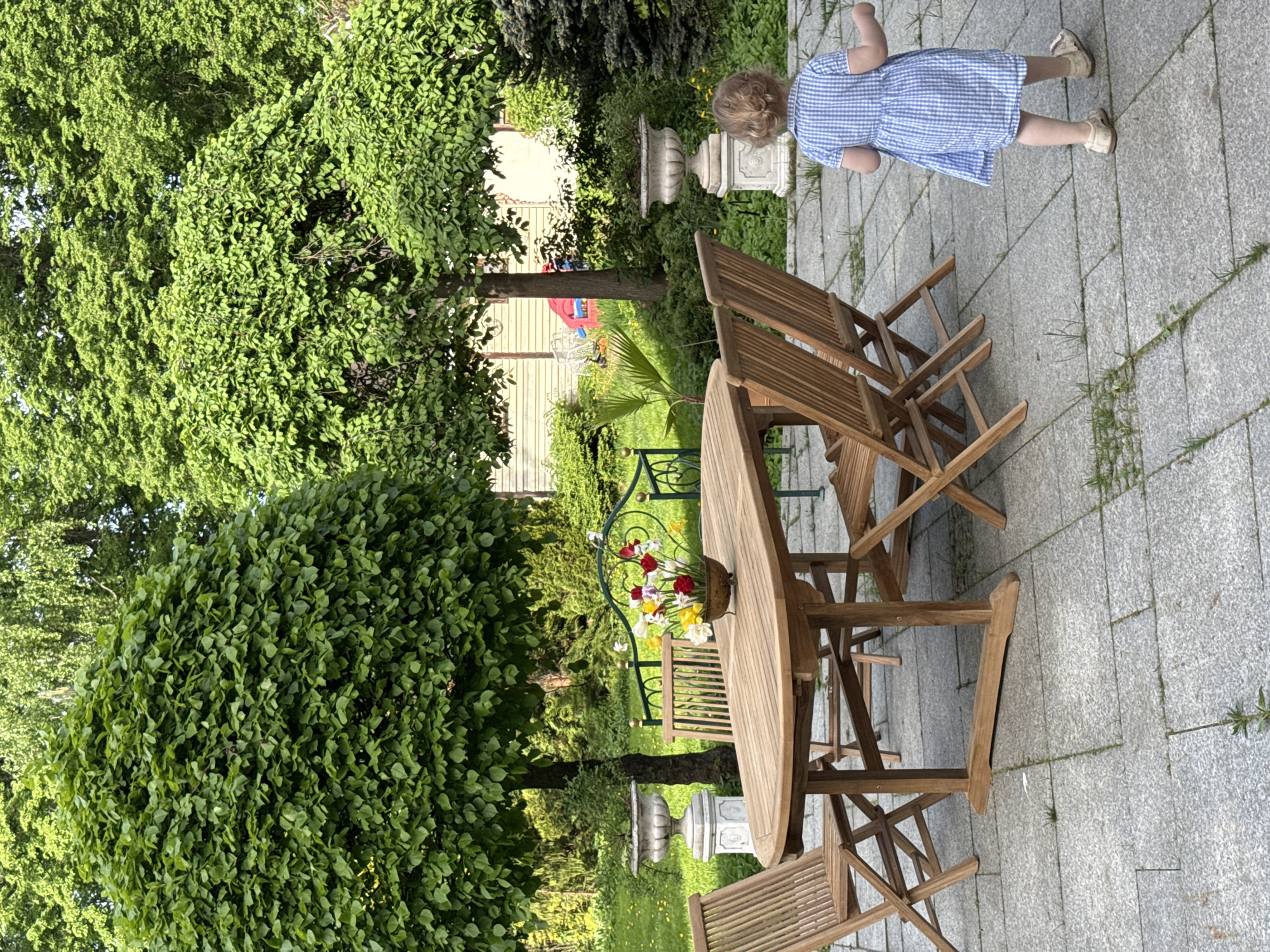 The image captures a garden scene in Sapronovo, Russia. A young child in a blue and white checked dress stands on a gray-paved patio. In the center of the image is a wooden table surrounded by wooden folding chairs, with a vase of flowers on the table. Lush green trees and bushes frame the scene. In the background, elements of a house are visible. A stone urn is positioned near the child.