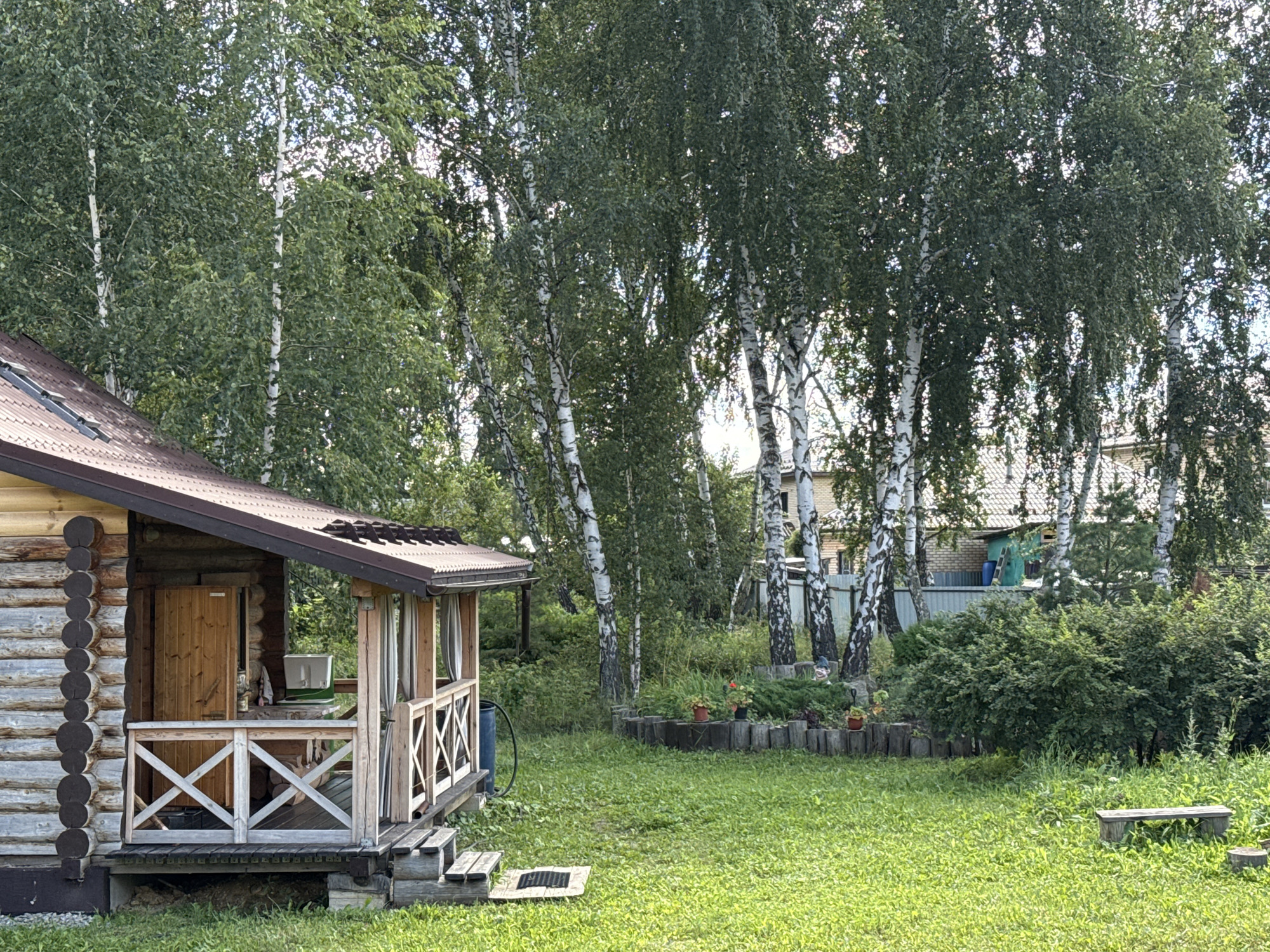 The image depicts a rustic wooden house with a covered porch, situated in a natural, grassy environment with numerous birch trees. The house is constructed from logs and has a brown, corrugated roof. The porch features wooden railings with an X-shaped design, a wooden door, and is furnished with a white cooler. In the background, more birch trees with distinctive white bark are visible, along with parts of other houses and fences. The ground is covered in green grass, with some small potted plants and a wooden bench in the yard. The scene appears to be a residential property in a rural or semi-rural setting, possibly in the city of Kremenkul', Russia.