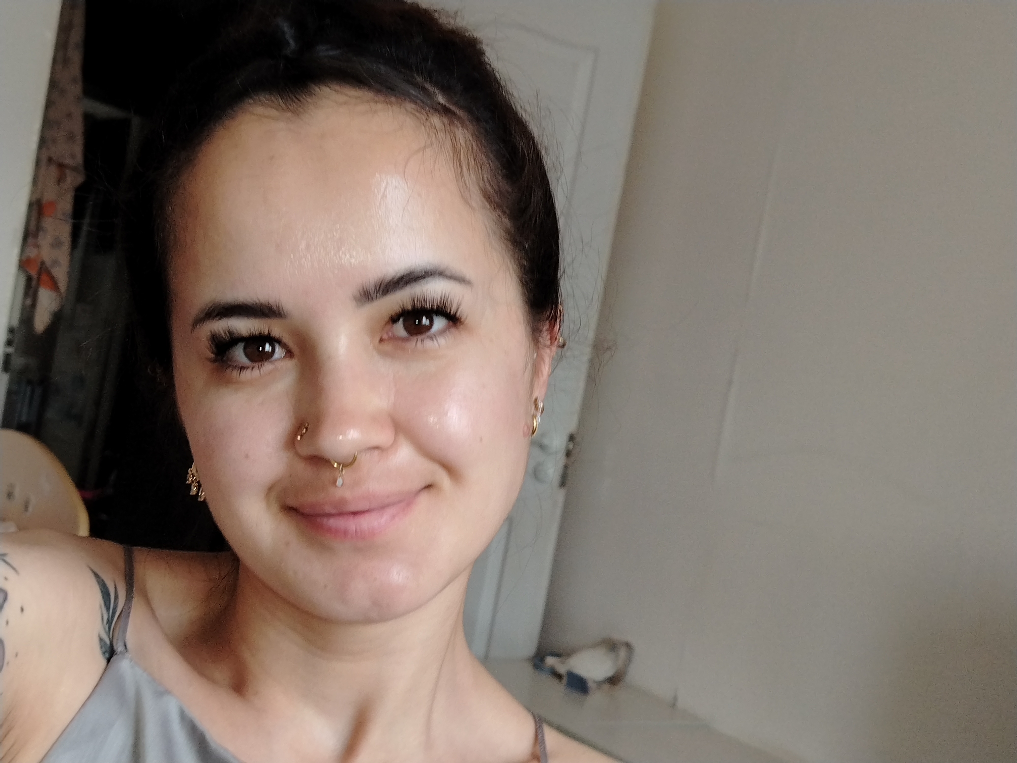 A close-up, chest-up, indoor selfie shows a young woman with her hair pulled up in a bun. She is wearing a grey tank top and has multiple piercings, including a septum ring and earrings. The background is a plain wall. The location in the city of Noviy, Russia, is not discernible from the image.