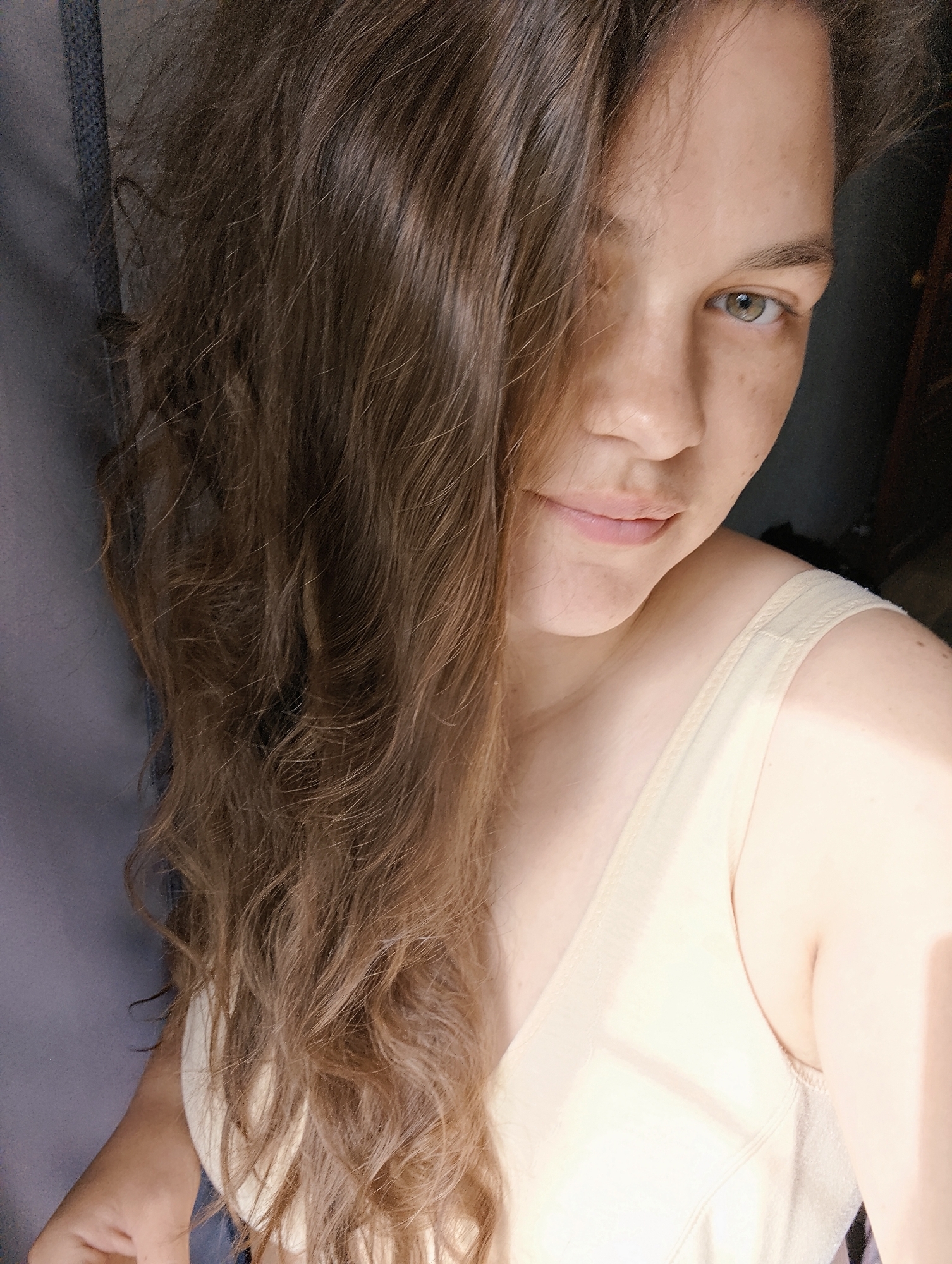 A medium-shot selfie shows a woman with long, wavy brown hair, green eyes, and fair skin. Part of her hair covers the left side of her face. She is wearing a light-colored sleeveless top. The background includes a gray curtain or wall on the left and a dark space on the right. The location in the city of Chaikovsky, Russia, cannot be determined from the image.