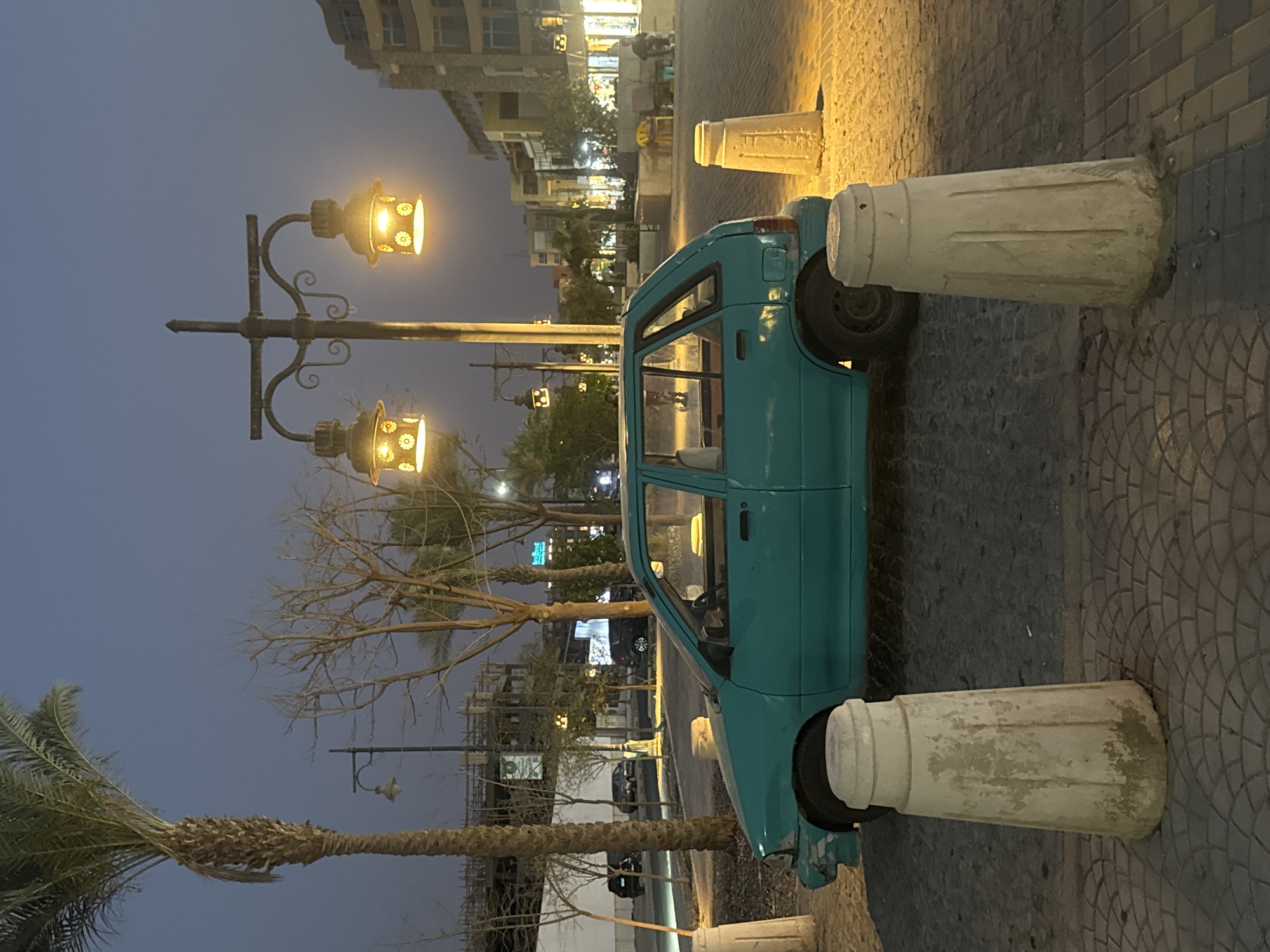 The image depicts a teal-colored car parked between two concrete bollards on a brick-paved street in an urban environment at night. Overhead street lamps illuminate the scene, creating pools of light. In the background, buildings line the street, with faint lights indicating activity. A palm tree is visible in the foreground, adding a tropical element to the scene.