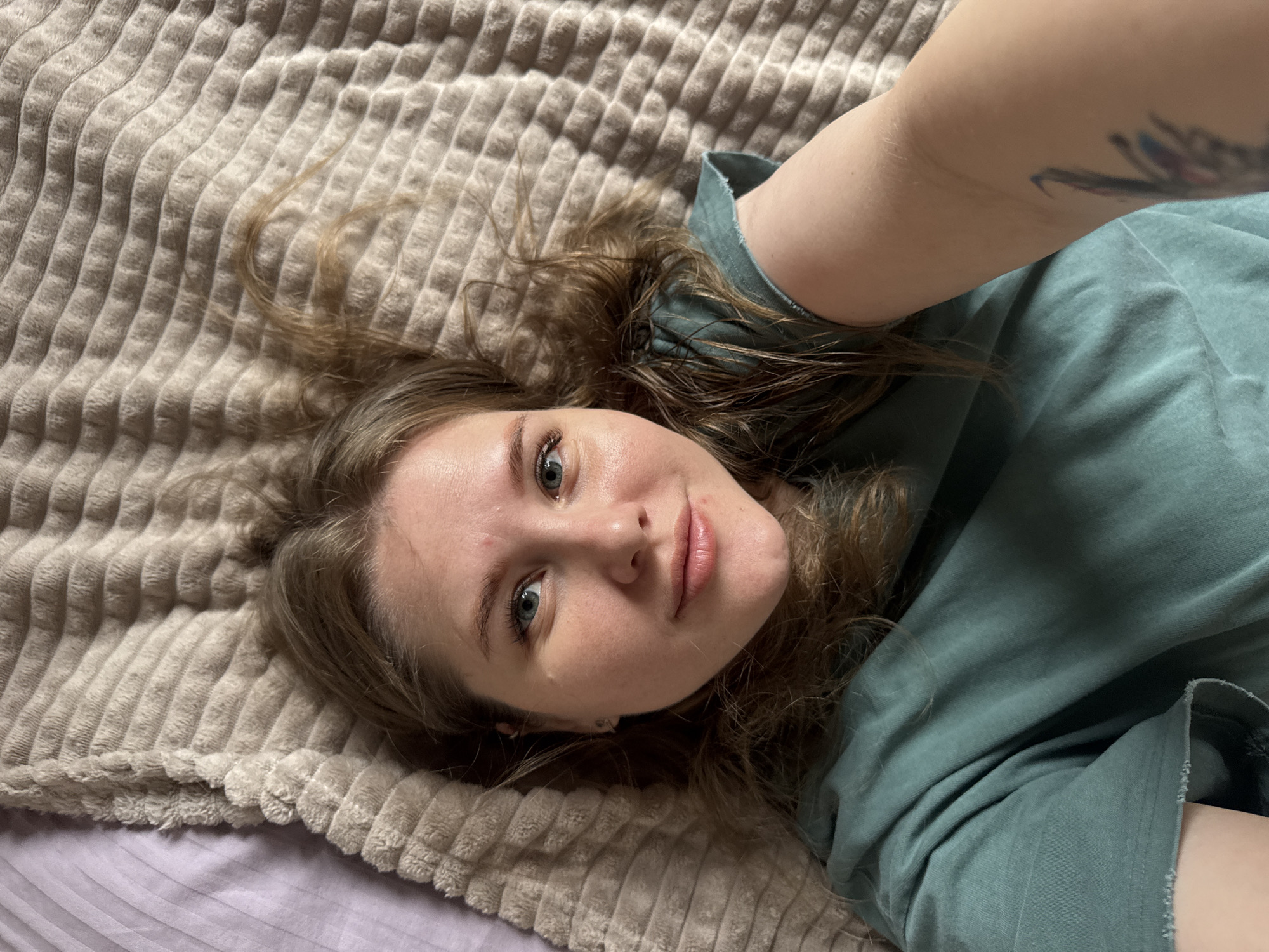A person is lying on a bed. The person has fair skin, light brown hair, and is wearing a green shirt. A portion of the person's arm with a tattoo is visible. The bed has a textured brown blanket and a lilac sheet underneath. The location is unspecified and cannot be determined from the image.