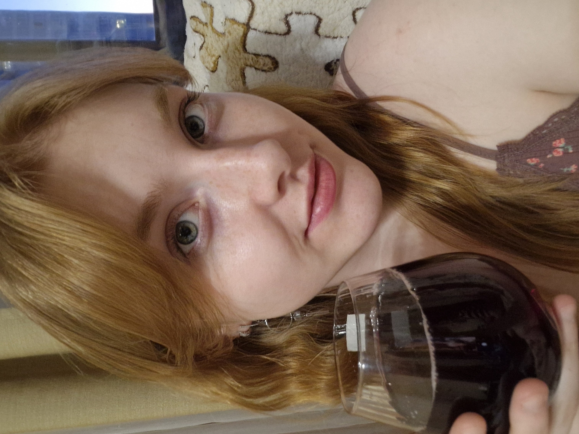 The image captures a close-up portrait of a young woman lying down indoors in Yekaterinburg, Russia, holding a glass of dark red liquid, likely wine. The photo is tilted, presenting the woman on her side. She has light skin with subtle freckles, long, flowing light red or auburn hair, and striking green eyes that engage directly with the camera. Her lips are a natural light pink. She is wearing a dark brown or maroon top with thin straps, featuring a delicate floral pattern with small pink blossoms visible on the fabric.
Behind her head is a cream-colored cushion with a distinctive brown puzzle-piece pattern. A window is visible in the upper left background, showing a soft blue light, suggesting it is daytime. The lighting is natural and gentle. The woman's expression is calm and neutral. Her hand gently grips the stem and base of the wine glass. The scene conveys a relaxed, informal setting. There is no visible text in the image.