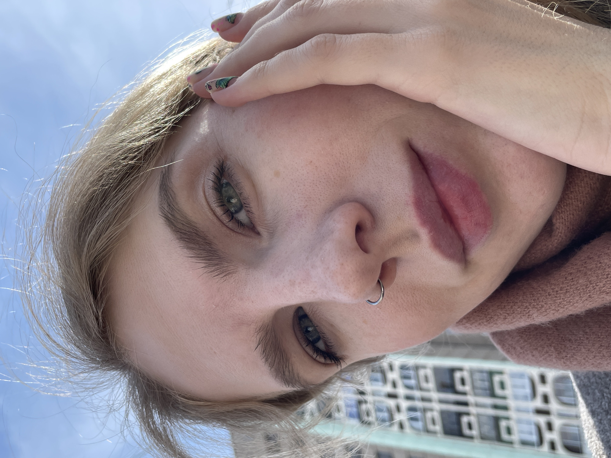 A close-up, rotated view of a fair-skinned person with light blonde hair and green eyes, partially obscuring their face with their hand. The fingers have colorful nail art. The person has a silver nose ring. They are wearing a pink sweater. The background includes a light blue sky and a building with a grid-like pattern. There is no information to discern if it is in Izhevsk, Russia.