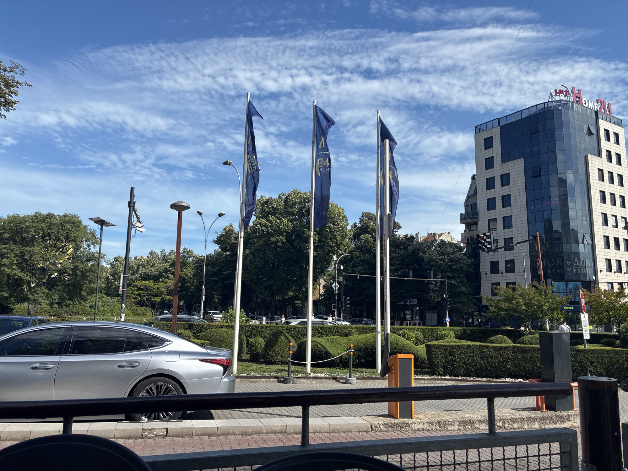 A medium shot captures a street scene in Varna, Bulgaria, on a bright, partly cloudy day. Three blue flags with yellow emblems, possibly advertising "HOME 24", are prominently displayed on poles in the foreground, slightly to the left of center. Behind the flags, a modern glass and concrete building with red lettering that reads "HOME 24 REAL ESTATE" rises into the sky. To the left of the building, a silver sedan is parked on the street. Lush green bushes and trees form a manicured landscape in the mid-ground, with a distinct topiary sculpture of a reclining figure visible. Streetlights and traffic signals are also present. The overall scene depicts an urban environment with commercial buildings, landscaping, and a car, under a sky dotted with white, wispy clouds.