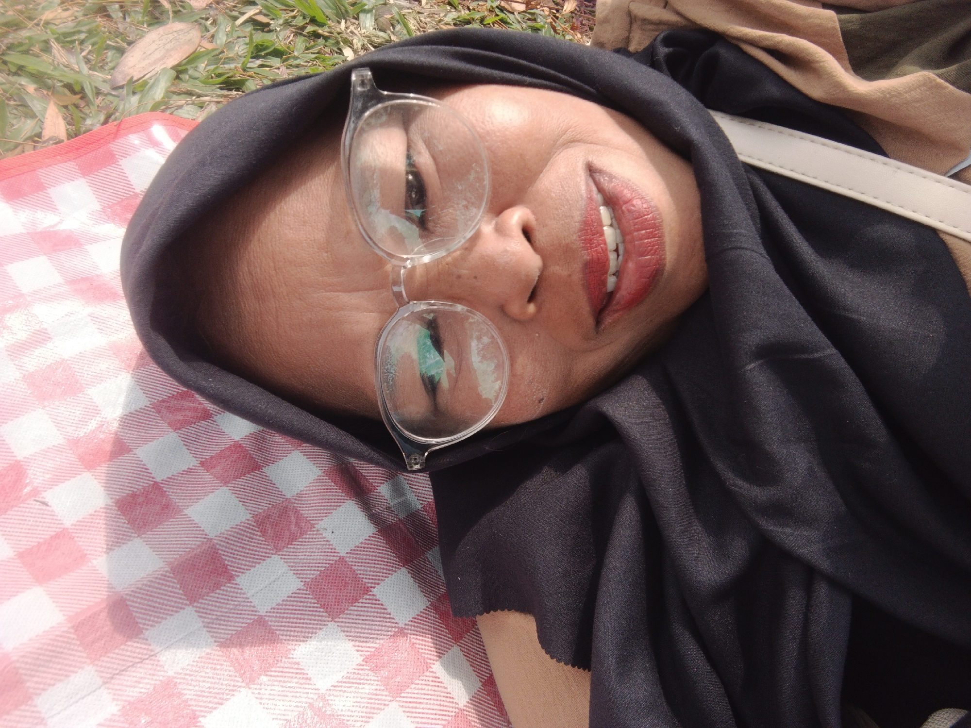 A woman wearing a black hijab and glasses lies on a pink and white checkered picnic blanket on grass. She is looking up at the camera with her mouth slightly open, revealing red lipstick and white teeth. A beige strap is visible across her chest. The background consists of dry grass and fallen leaves. The scene appears to be outdoors in Perbawati, Indonesia.